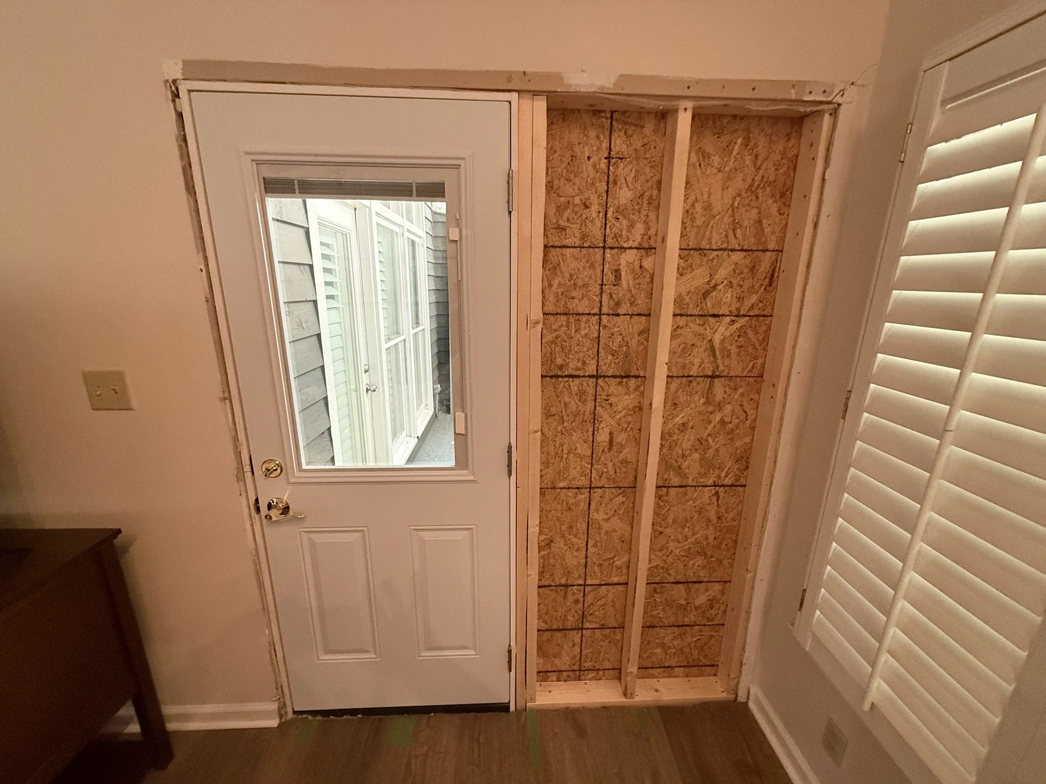 Interior view of a room with a white door and window, adjacent to a wall opening framed with wooden studs, with some drywall and a white plantation shutter.