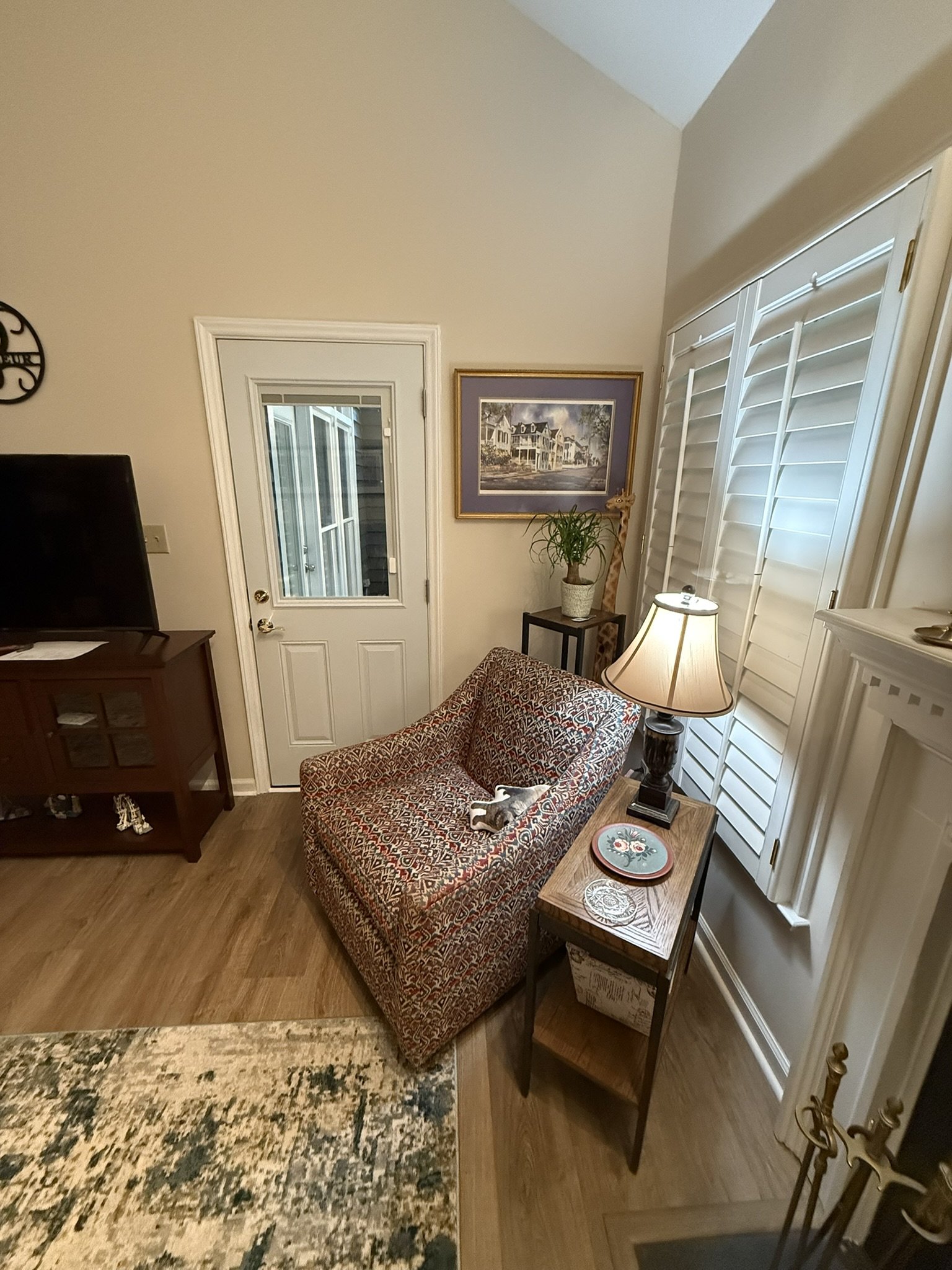 Living room with a patterned couch, a wooden side table with decorative plates, a lamp, a framed painting of houses, a tall potted plant, and a wall-mounted clock. There are windows with white shutters and an exterior door with a glass panel.