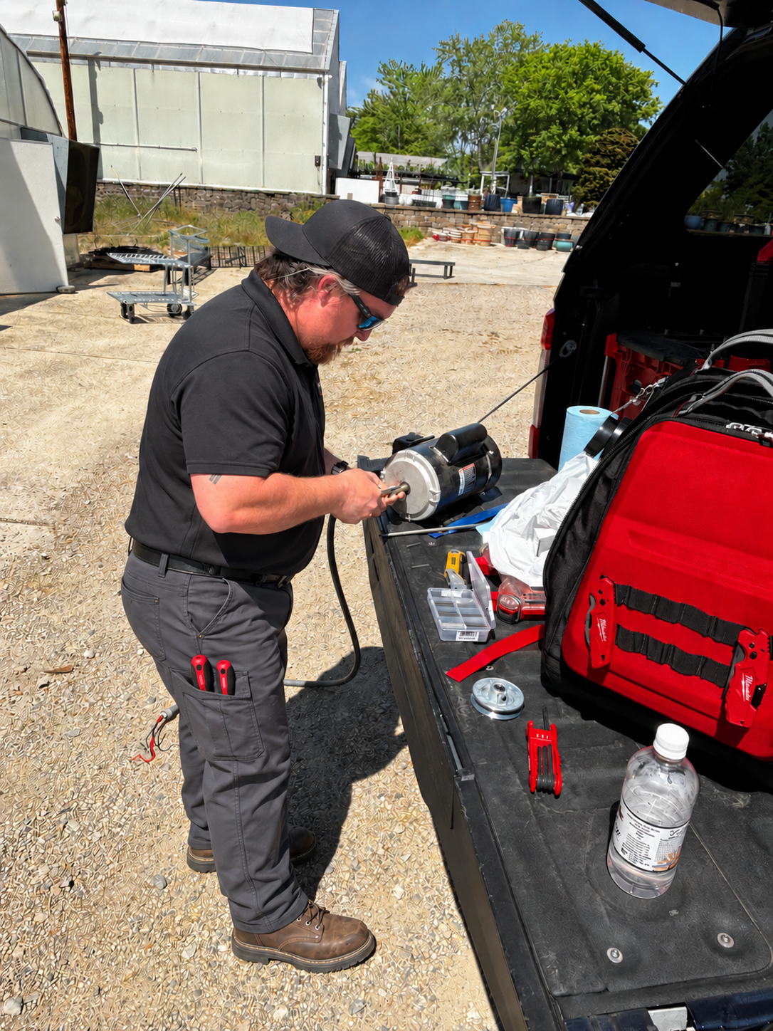 A man working on a Motor at a work table in an outdoor setting with green trees, potted plants, and a greenhouse in the background.