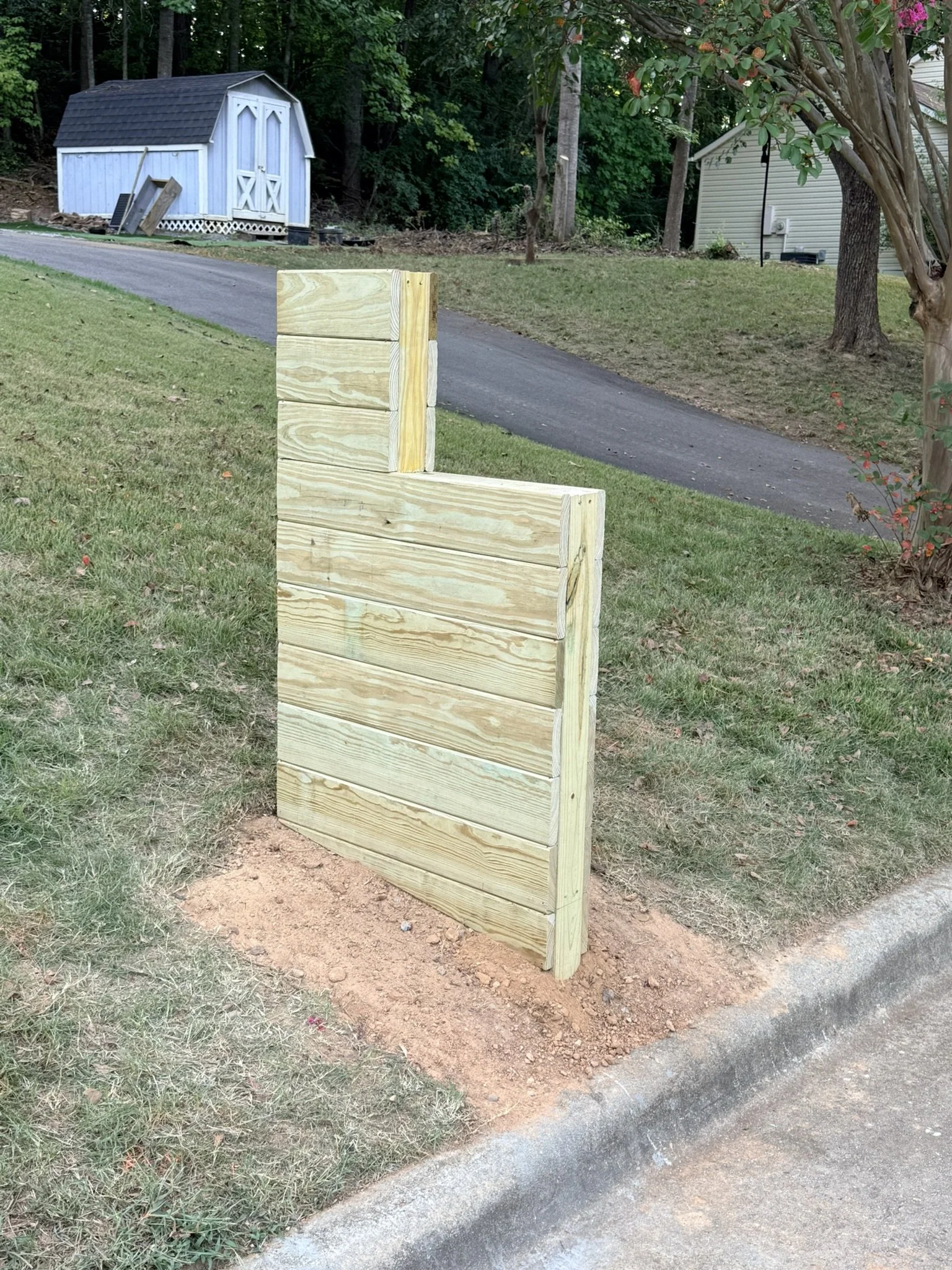 A yard with a partially finished wooden mailbox near a curb, with a paved driveway, trees, and two small sheds in the background.