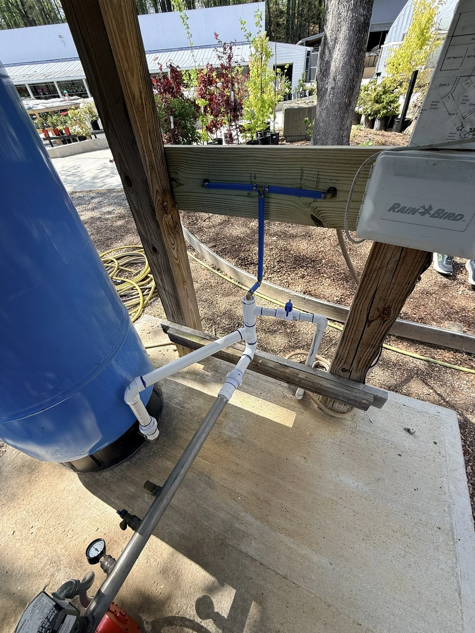 A water filtration system setup outdoors on a concrete slab, including a blue water storage tank, PVC pipes with valves and a pressure gauge, and a Rain Bird control box mounted on wooden supports.