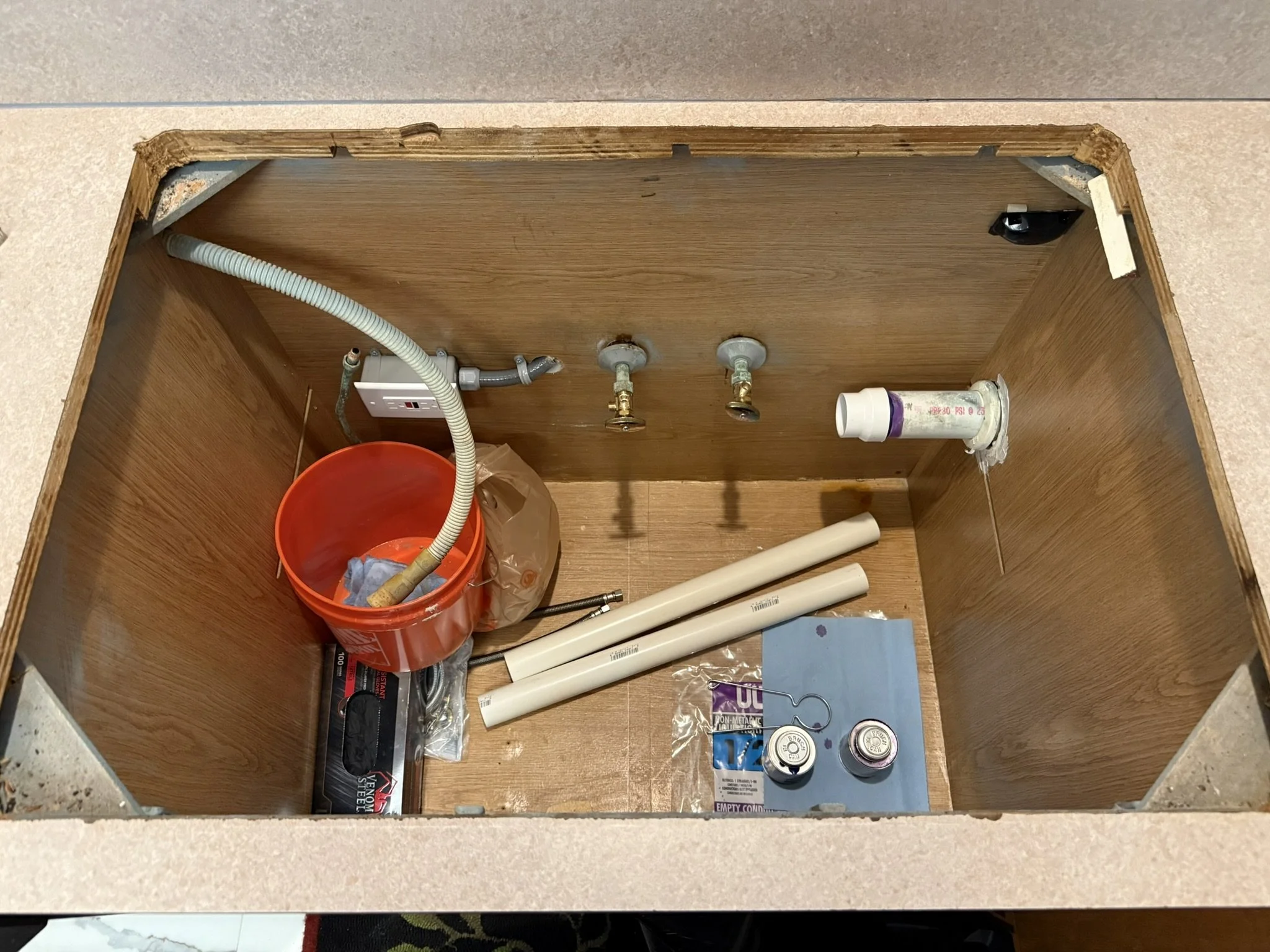 Inside the cabinet, there are plumbing pipes, a red bucket, tools, and various plumbing supplies for installation or repair.