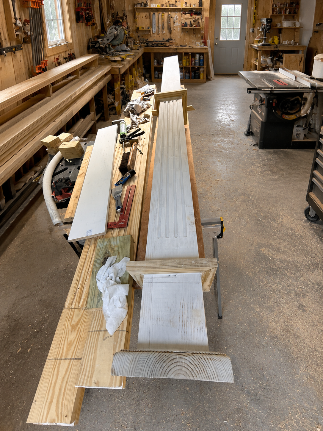 Woodworking workshop with a long workbench, various woodworking tools, and materials, including a metal piece, on the bench; shelves with supplies and a saw machine in the background.