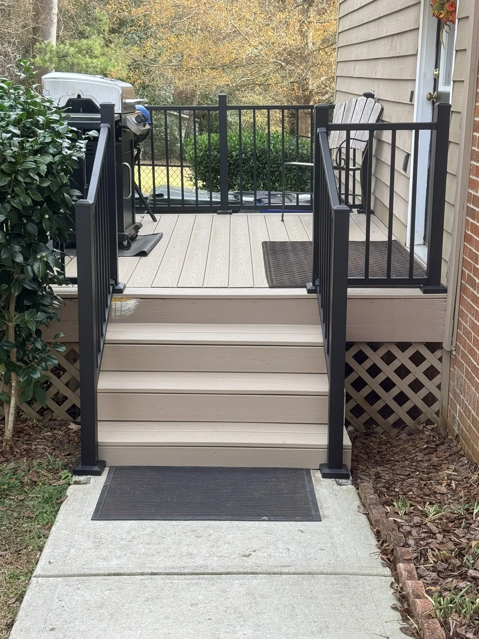 Small porch with beige Trex steps, black metal railing, outdoor grill, black doormat, side of house with beige siding, small potted plant on right, garden with mulch on left, trees in background.