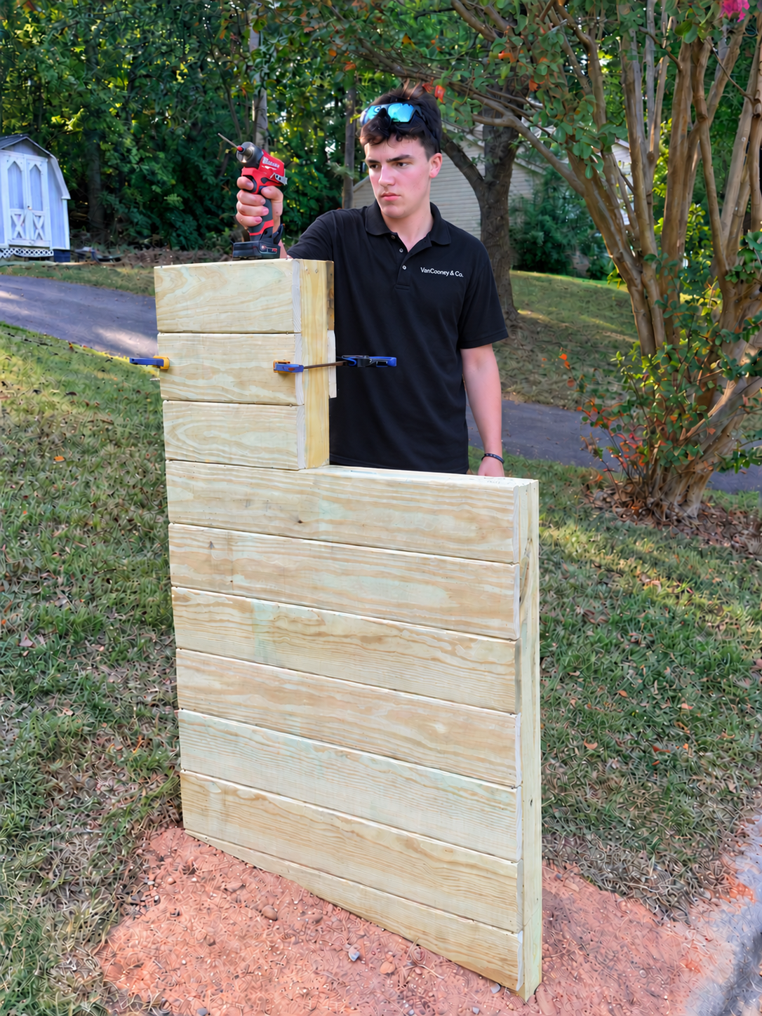A young man stands outdoors, holding a cordless drill, working on a wooden mailbox with horizontal boards, while clamped to a larger wooden structure, in a green backyard with trees and garden plants.