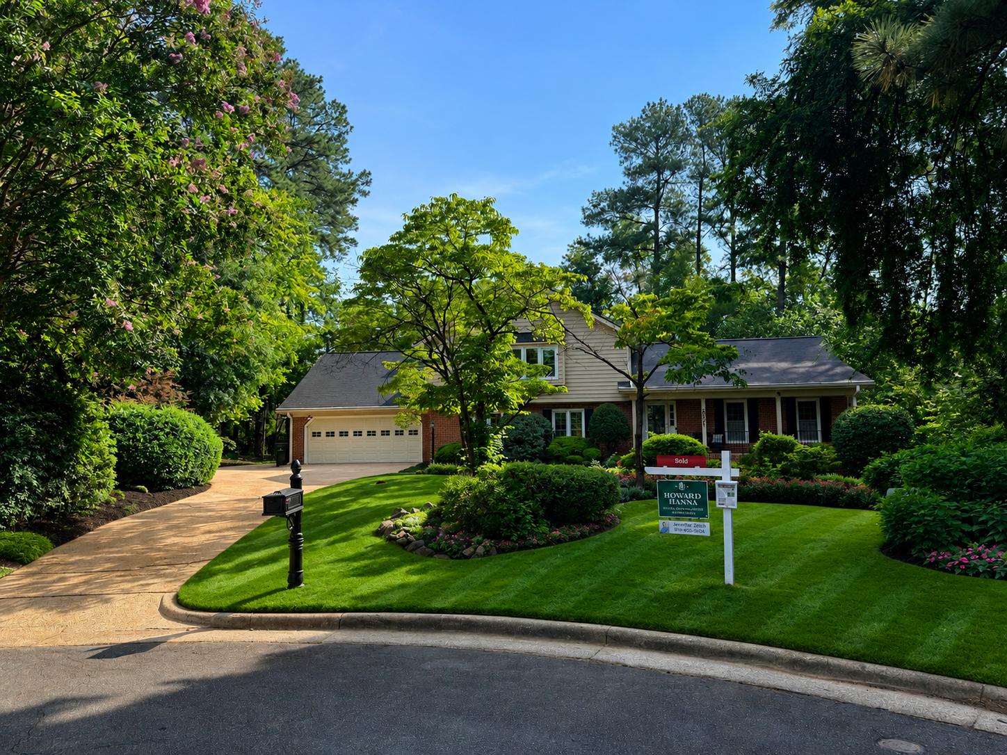 A well-maintained front yard with lush green grass, flowering bushes, and small trees in front of a house with a two-car garage. There is a black mailbox on a post near the sidewalk, and signs indicating the house is sold and for sale by a real estate company.