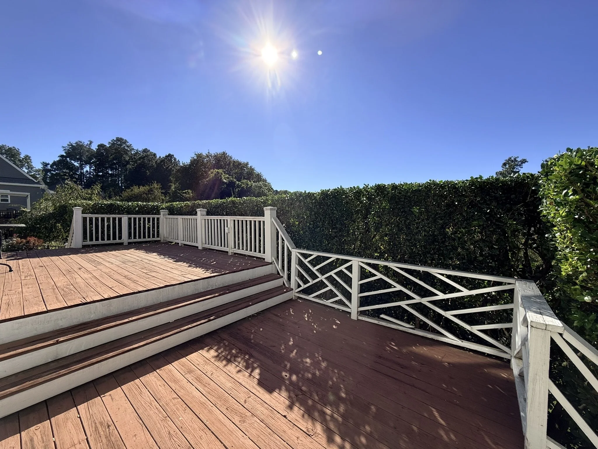 A wooden deck with white railing and stairs, surrounded by green bushes and trees, under a clear blue sky with the sun shining brightly.