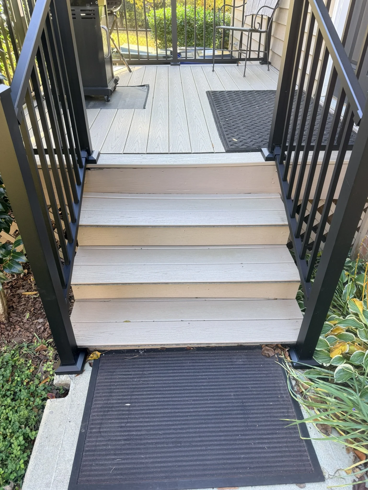 A small porch with a black metal railing, a black doormat, a grill, a metal chair, and some plants and bushes around the edges.
