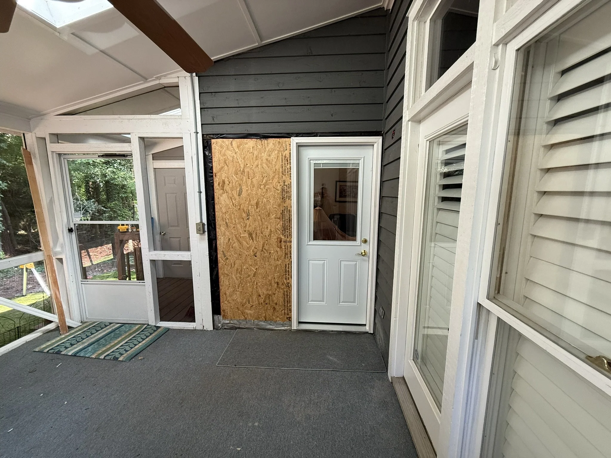 An enclosed porch or sunroom with large windows and sliding glass doors, a door with a window leading outside, black siding on the upper wall, and a partially installed wall covered with plywood. There is a small striped rug on gray carpeting, and th