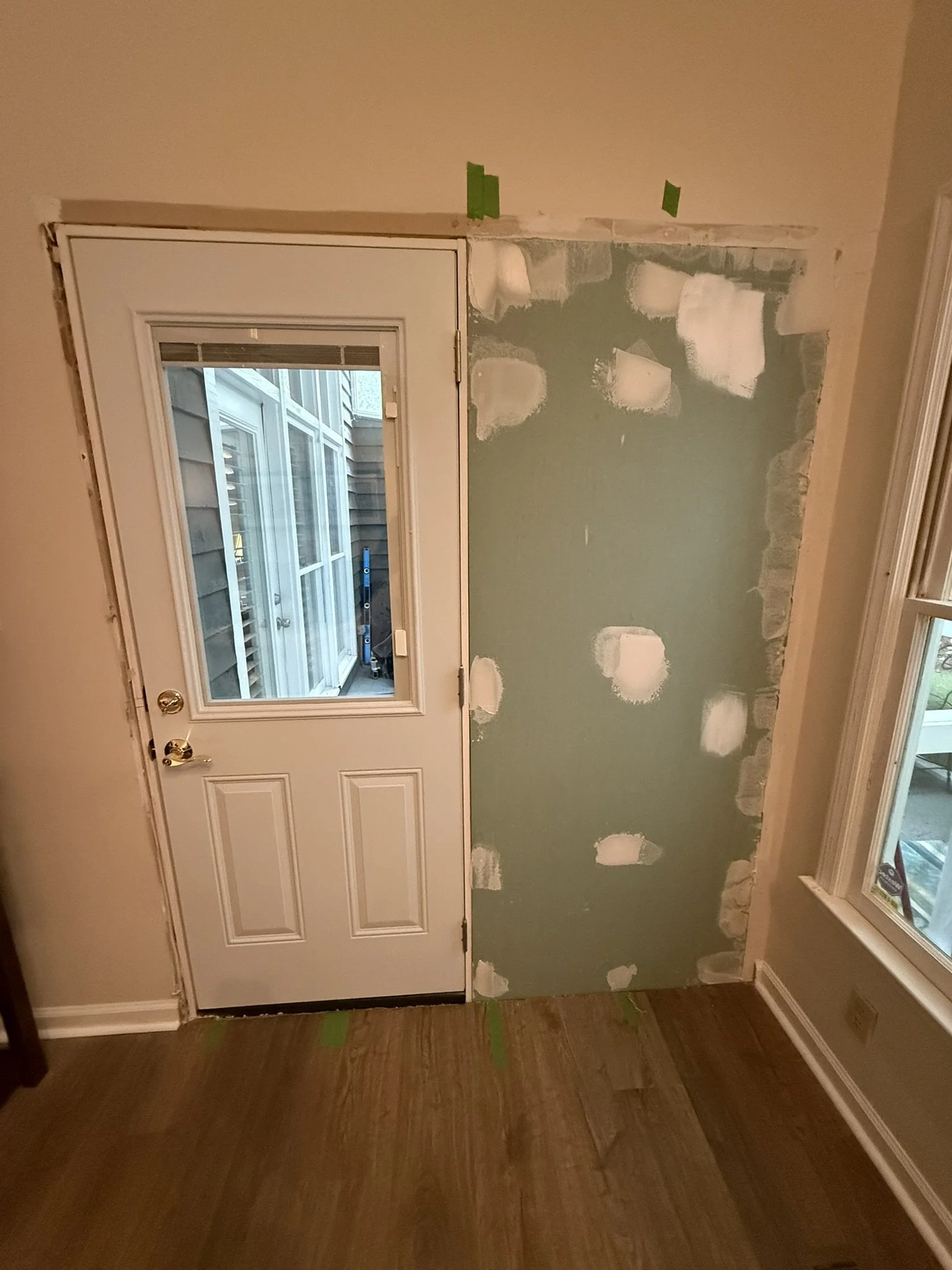 Interior door with window leading to a porch or yard, part of a wall under renovation with drywall patches and green painter's tape, hardwood flooring, and a window to the side.