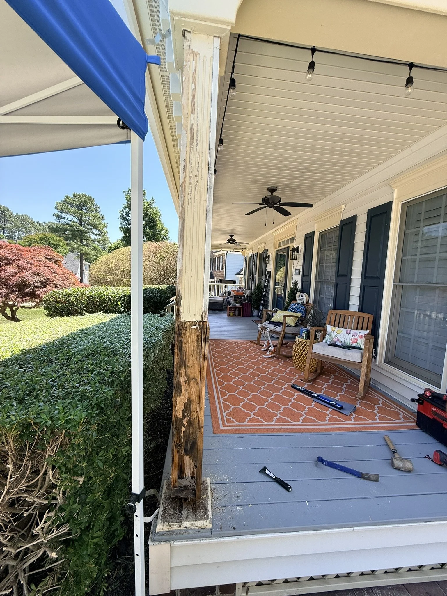 The porch of a house is under renovation, with tools scattered on the floor, chairs, and a rug. The porch has ceiling fans and string lights, with a view of trees and bushes outside.
