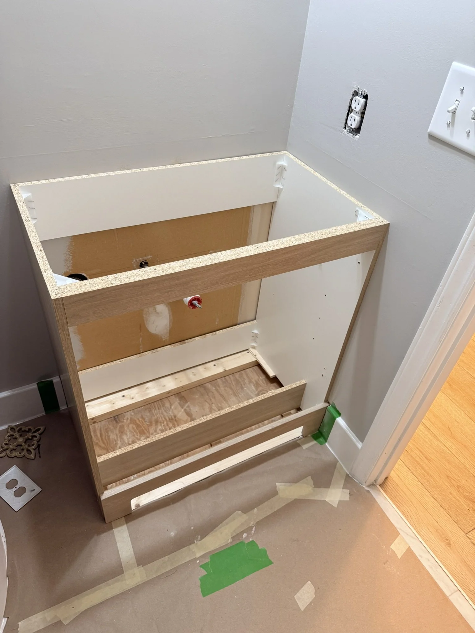 A partially assembled bathroom vanity with an open top, wooden frame, and pipes visible underneath, next to electrical outlets and wall switches on a wall.