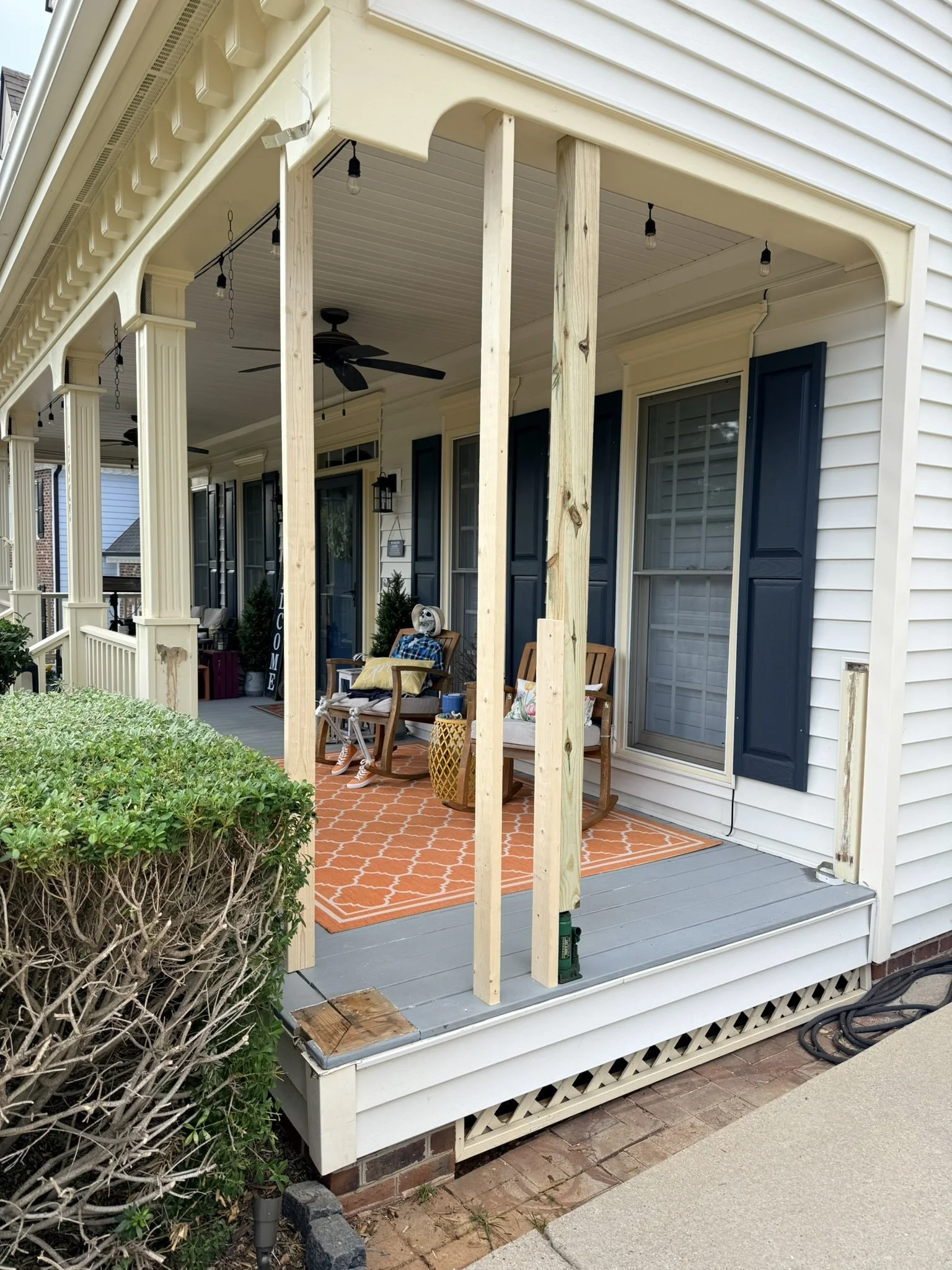 The image shows a house porch undergoing renovation. The porch has a new ceiling fan, black light fixtures, and string lights. It is partially enclosed with vertical posts, with some parts still under construction. There are two chairs with cushions 
