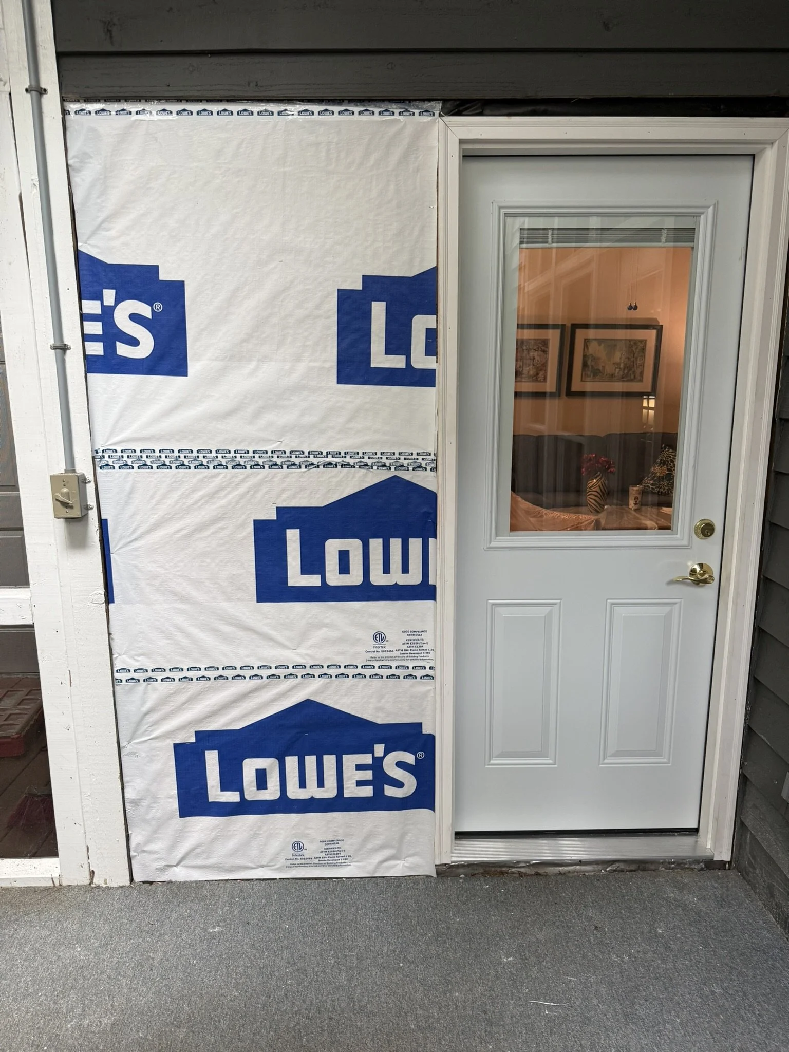 The exterior of a building with Lowe's branded house wrap on the left side and a white door on the right side, with a window in the door showing a living room with framed pictures, a lamp, and a decorative zebra-striped vase inside.