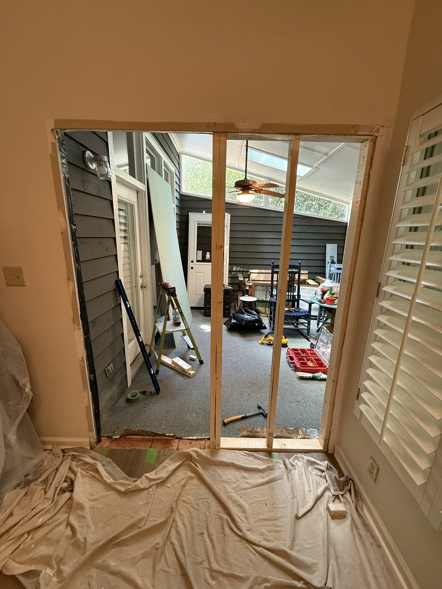 View of a room under renovation, showing a partially built wall frame with vertical studs, construction tools, and equipment, with an outdoor patio area visible through the opening.