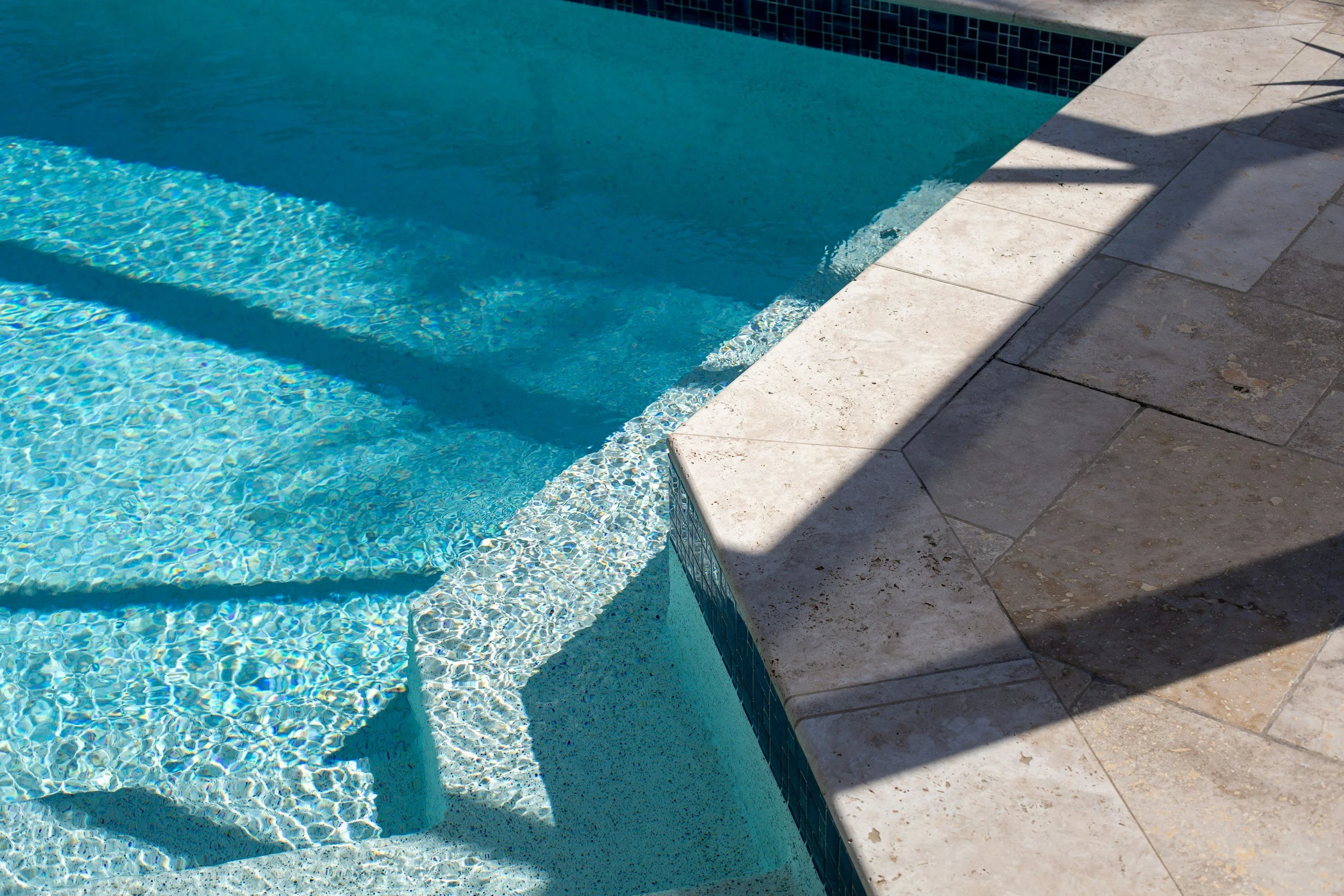A swimming pool with blue water and a tiled border, surrounded by a stone deck with shadows cast on it.