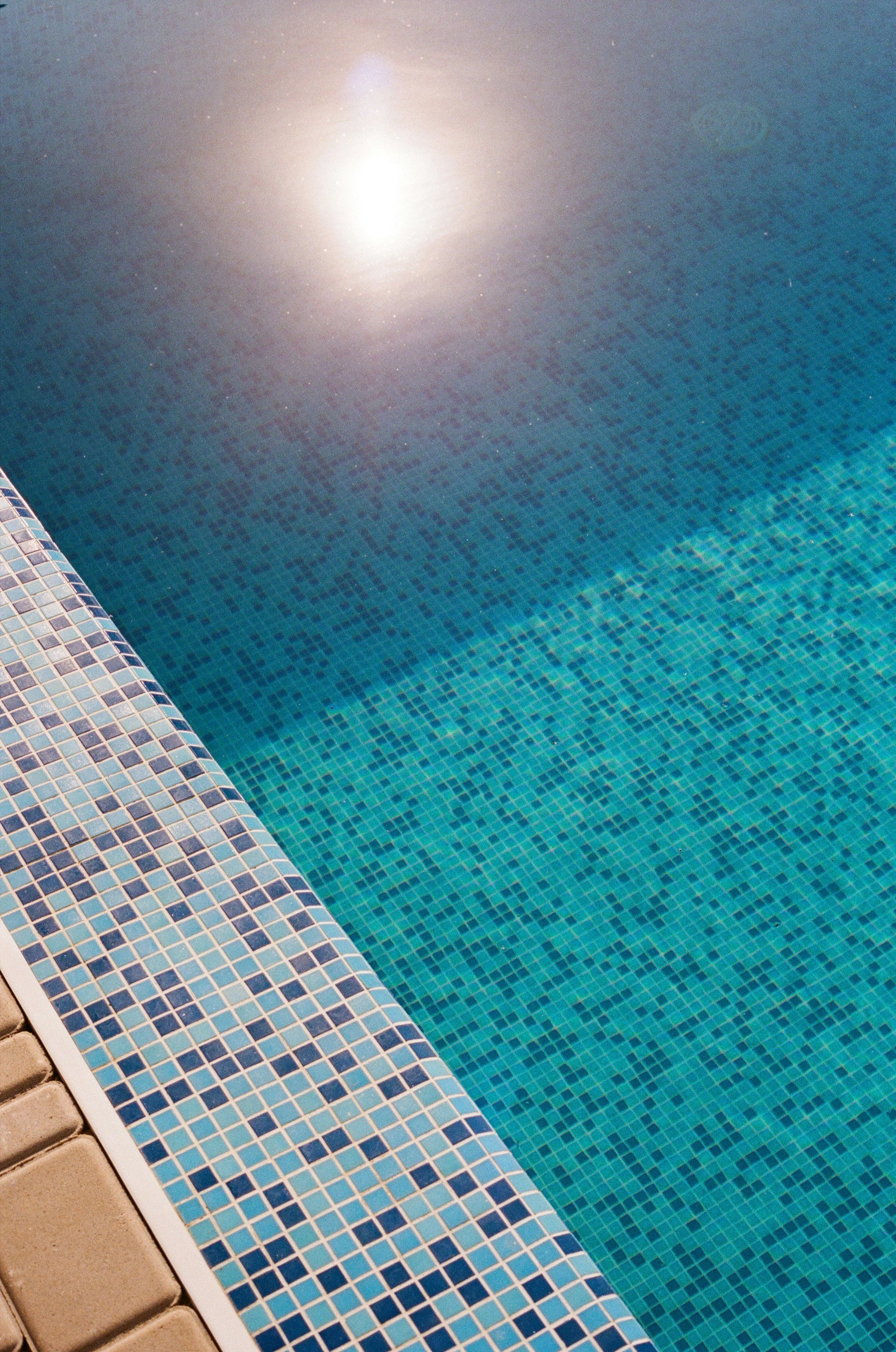Close-up of a swimming pool with blue mosaic tiles and a paved edge with small square tiles, reflecting sunlight.