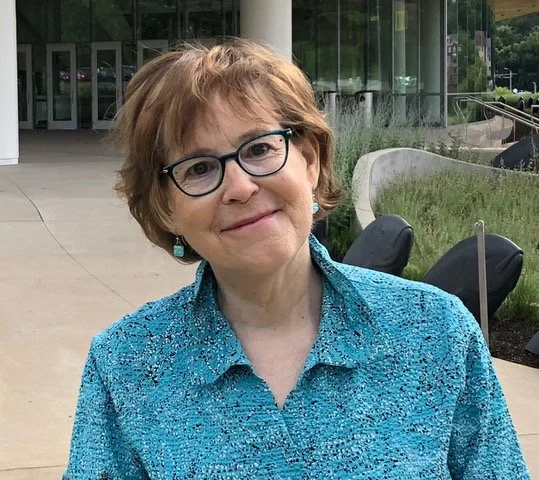 A portrait of children's author Tess Weaver. She smiles at the camera in black glasses and a blue patterned shirt, an outdoor public space behind her.