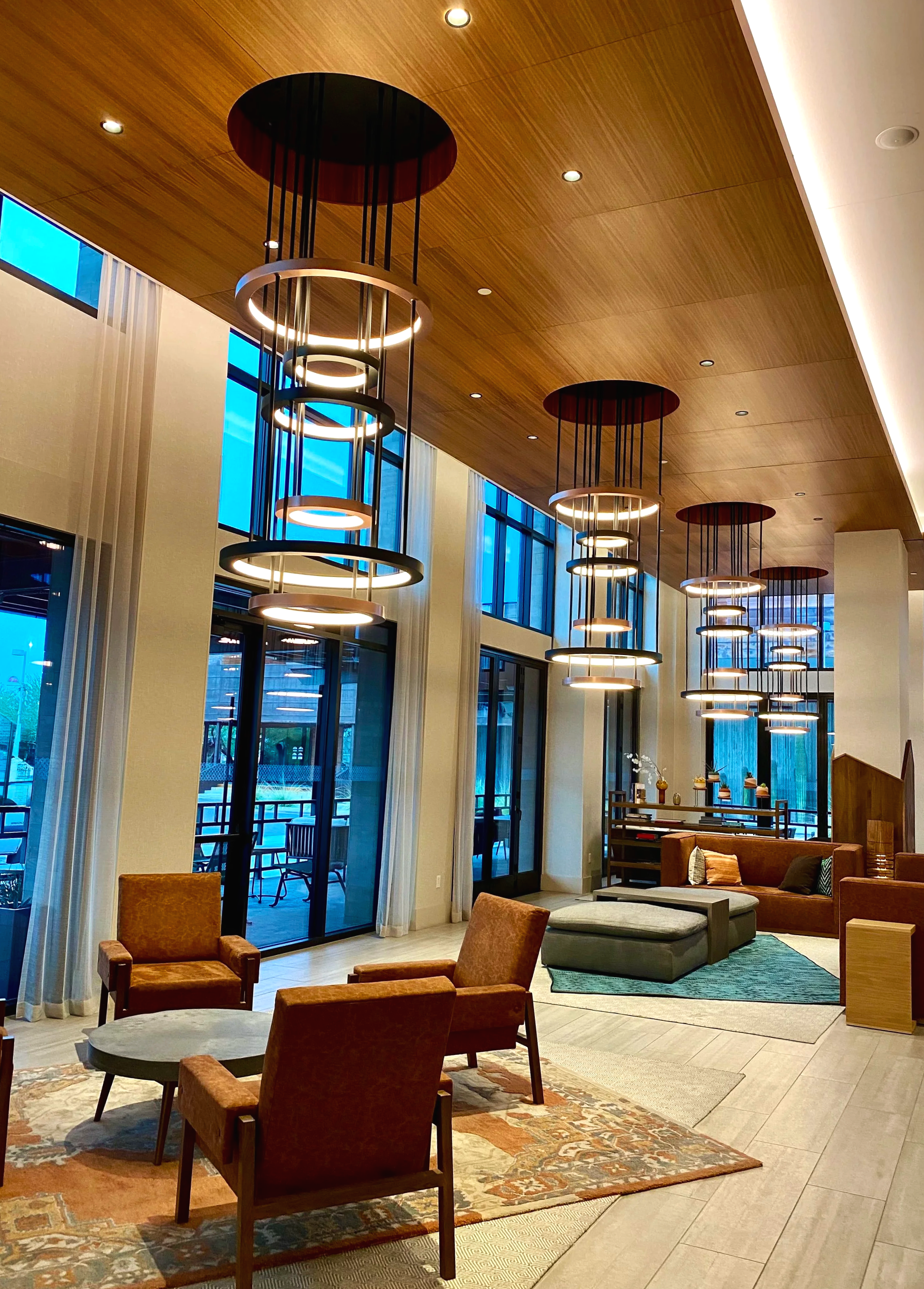 Interior of a modern hotel lobby with high ceiling, circular pendant lights, large windows, seating area with armchairs and sofas, decorative rug, and wooden accents.