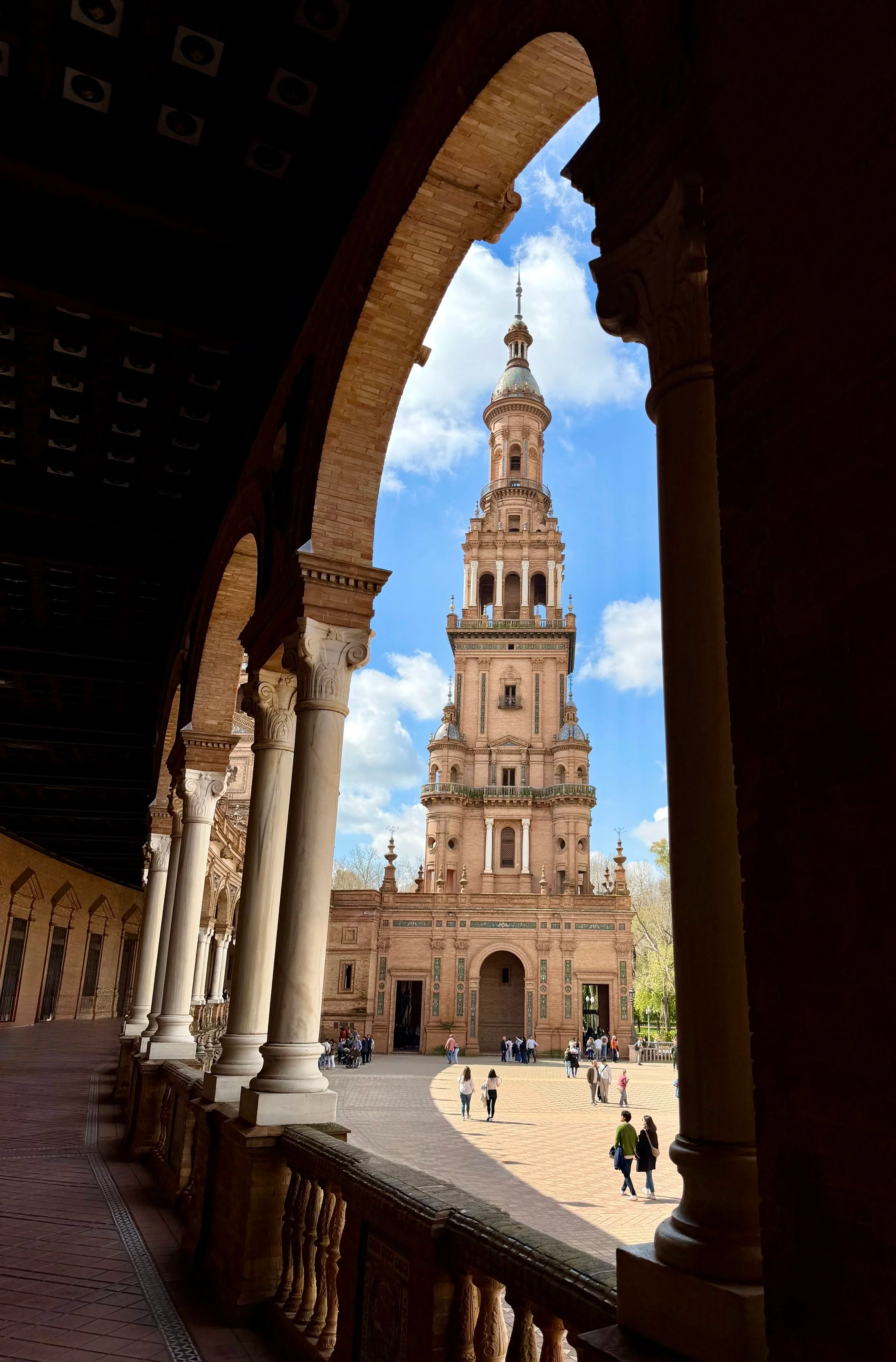 View of a historic tower, Plaza de España, through arches of a courtyard with columns, showing people walking in a sunny outdoor area.
