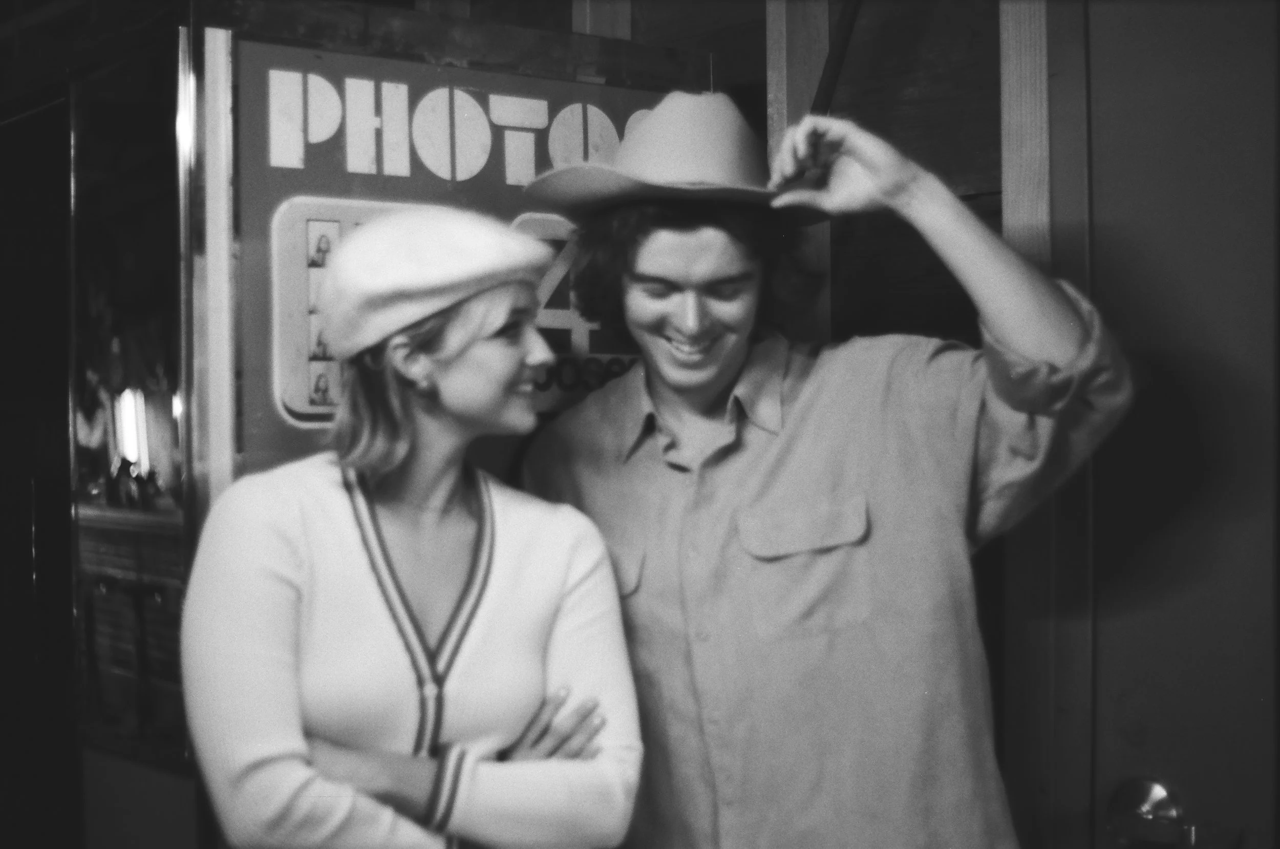 Black-and-white photo of two smiling people posing together indoors, with one adjusting the other's hat.
