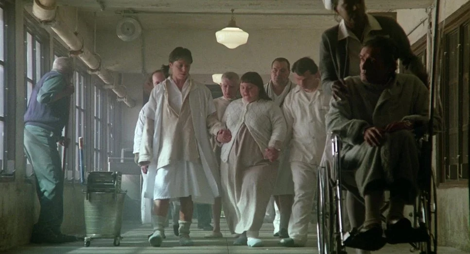 Group of psychiatric hospital patients and staff walking down an institutional corridor, with a person seated in a wheelchair.