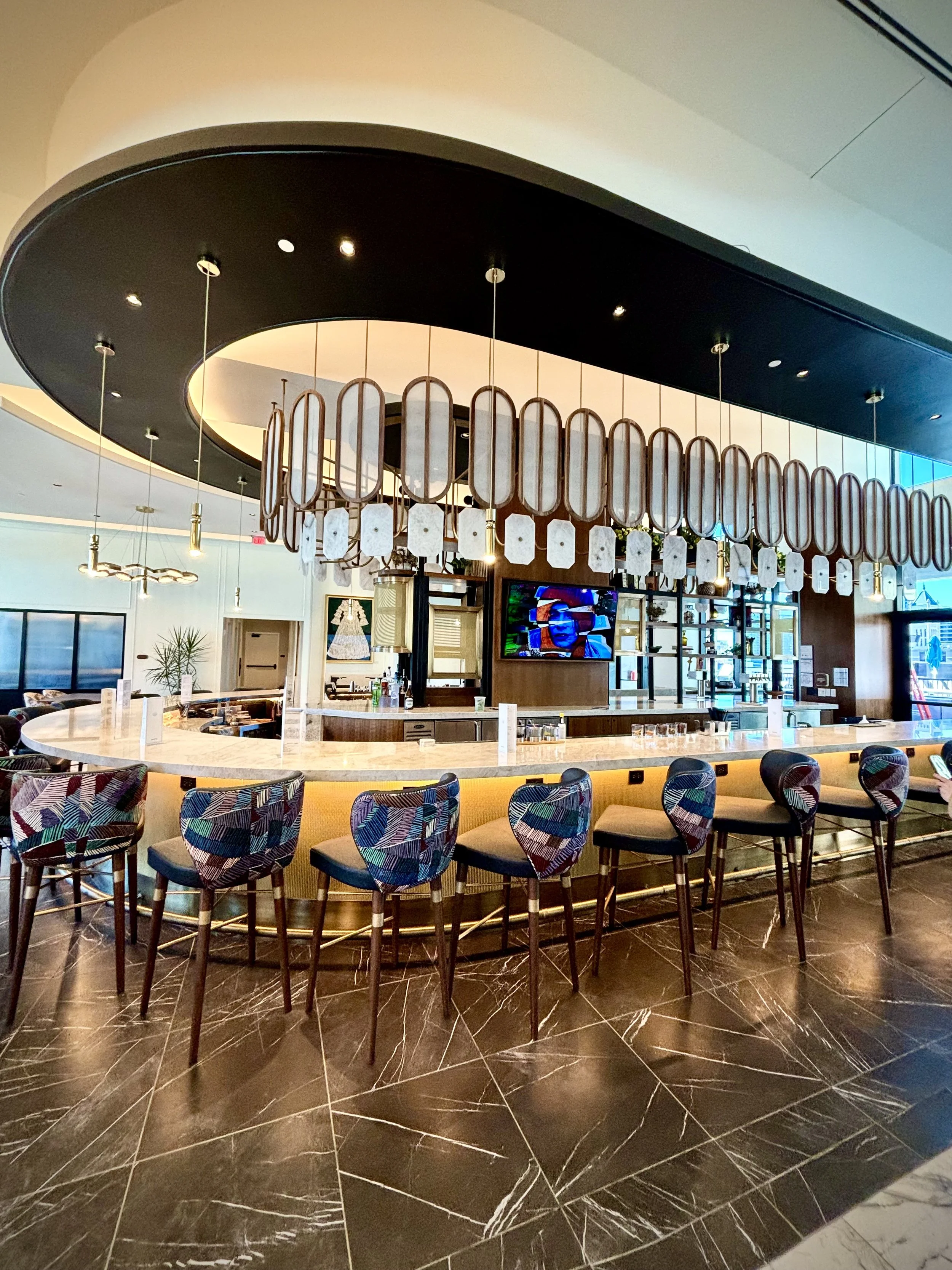 An upscale bar or restaurant interior featuring a curved counter with modern bar chairs with colorful, patterned fabric seats. Behind the bar, there is a decorative hanging light fixture with a black ceiling and hanging pendant lights. The floor is dark marble with white veining.