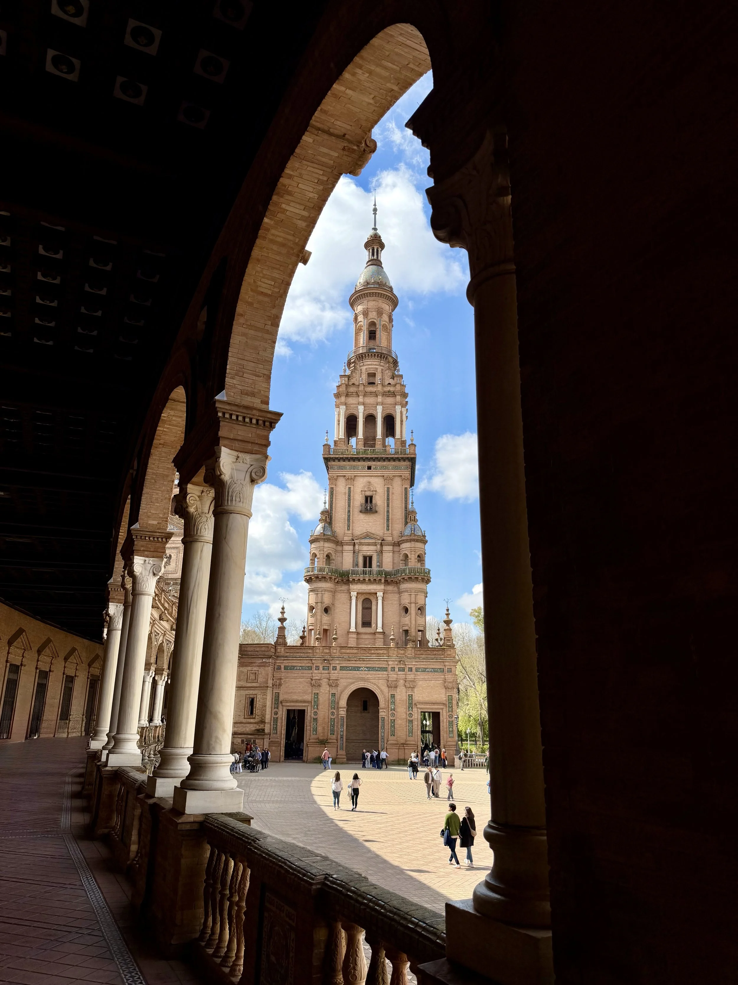 View of a tall historic tower with a spire, seen through arches and columns in a courtyard filled with tourists and visitors.