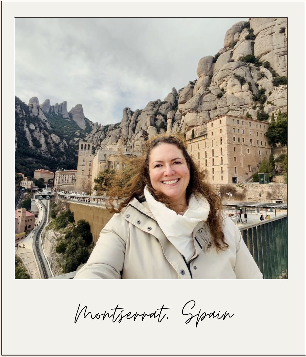 A woman with curly brown hair smiling in front of Montserrat, Spain, with rocky mountain formations and historic buildings in the background.