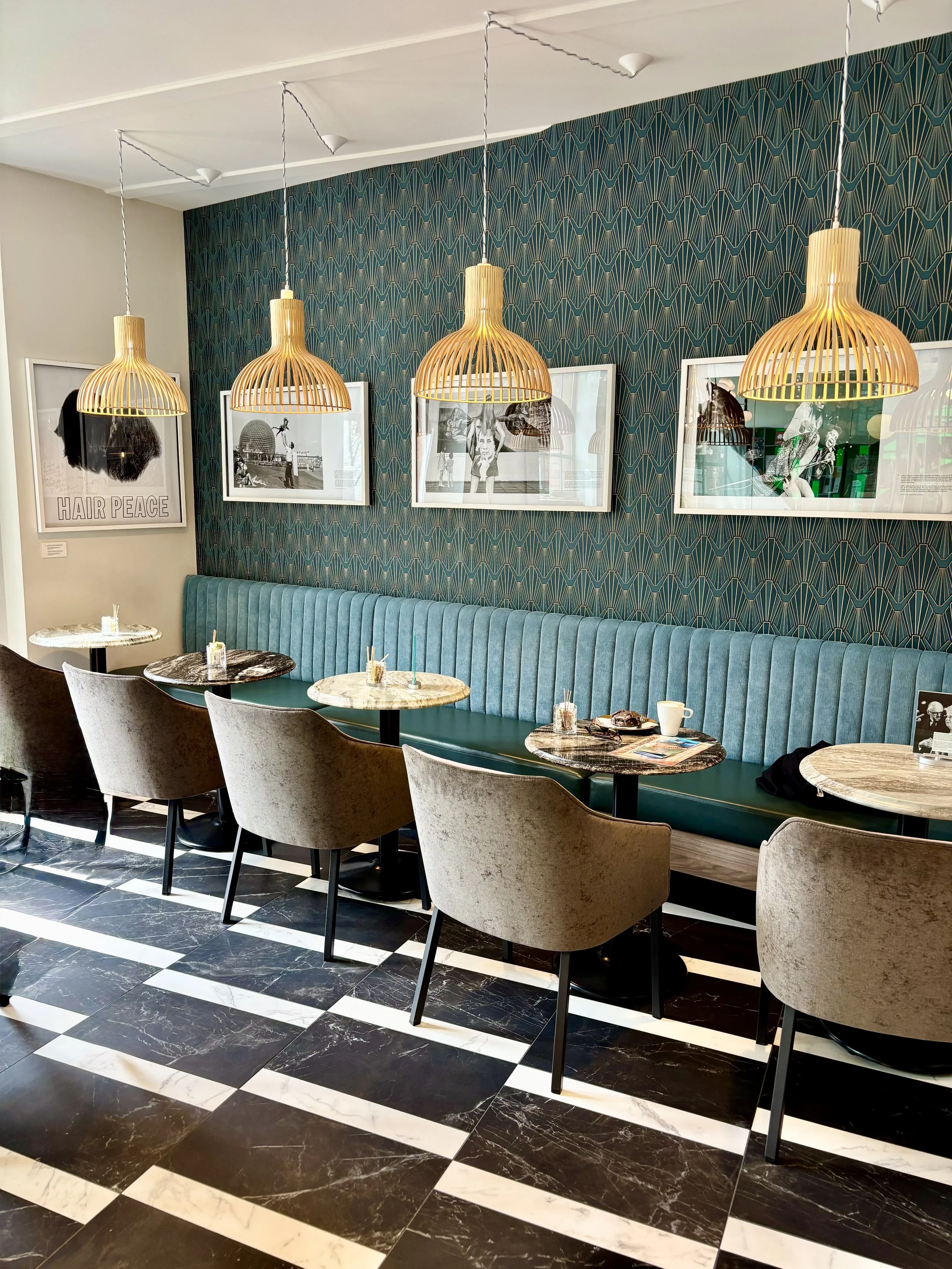 Interior of a cafe with a long teal cushioned banquette, four marble-top tables, and six beige chairs. Wall art includes framed black-and-white photos, and gold pendant lights hang from the ceiling. The floor is black and white checkered marble.
