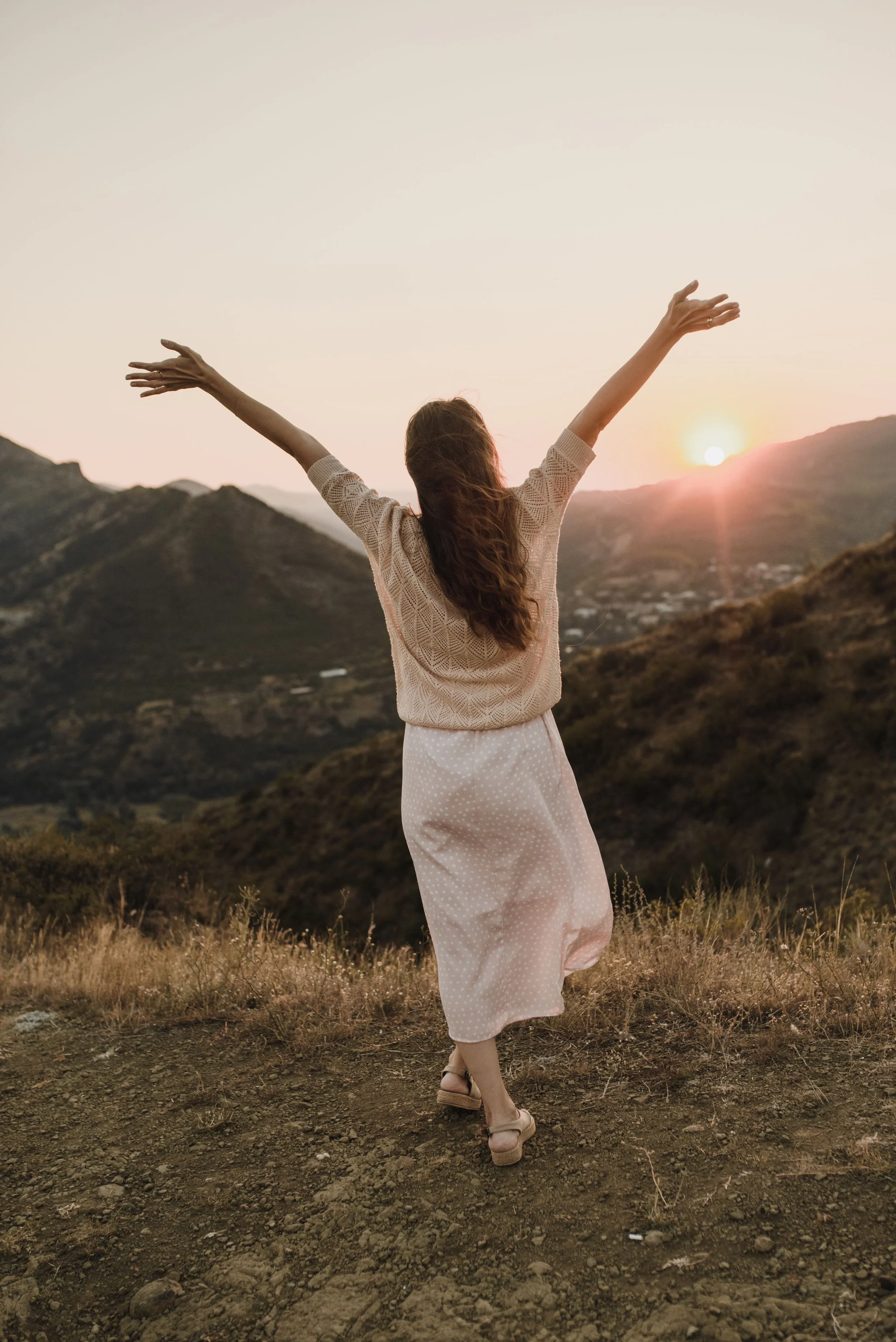 A woman with long hair wearing a beige sweater and a light-colored polka dot skirt standing in a field with mountains in the background during sunset, with arms raised toward the sky.