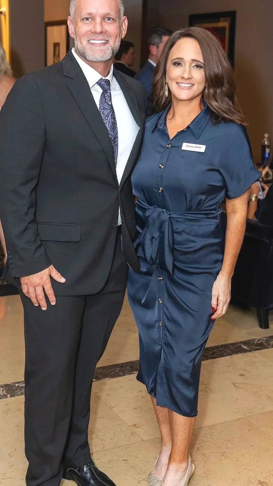 A man and a woman standing together, smiling at an indoor event. The woman is wearing a navy blue dress with a belt and has a name tag that says Casey Bolin. The man is dressed in a dark suit with a patterned tie.