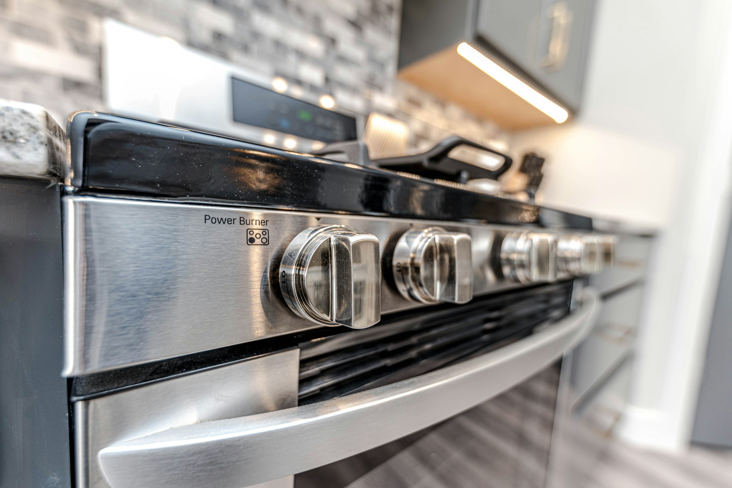 Close-up of a stainless steel gas stove with black control knobs, labeled 'Power Burner', in a modern kitchen with a brick backsplash and wooden cabinet.