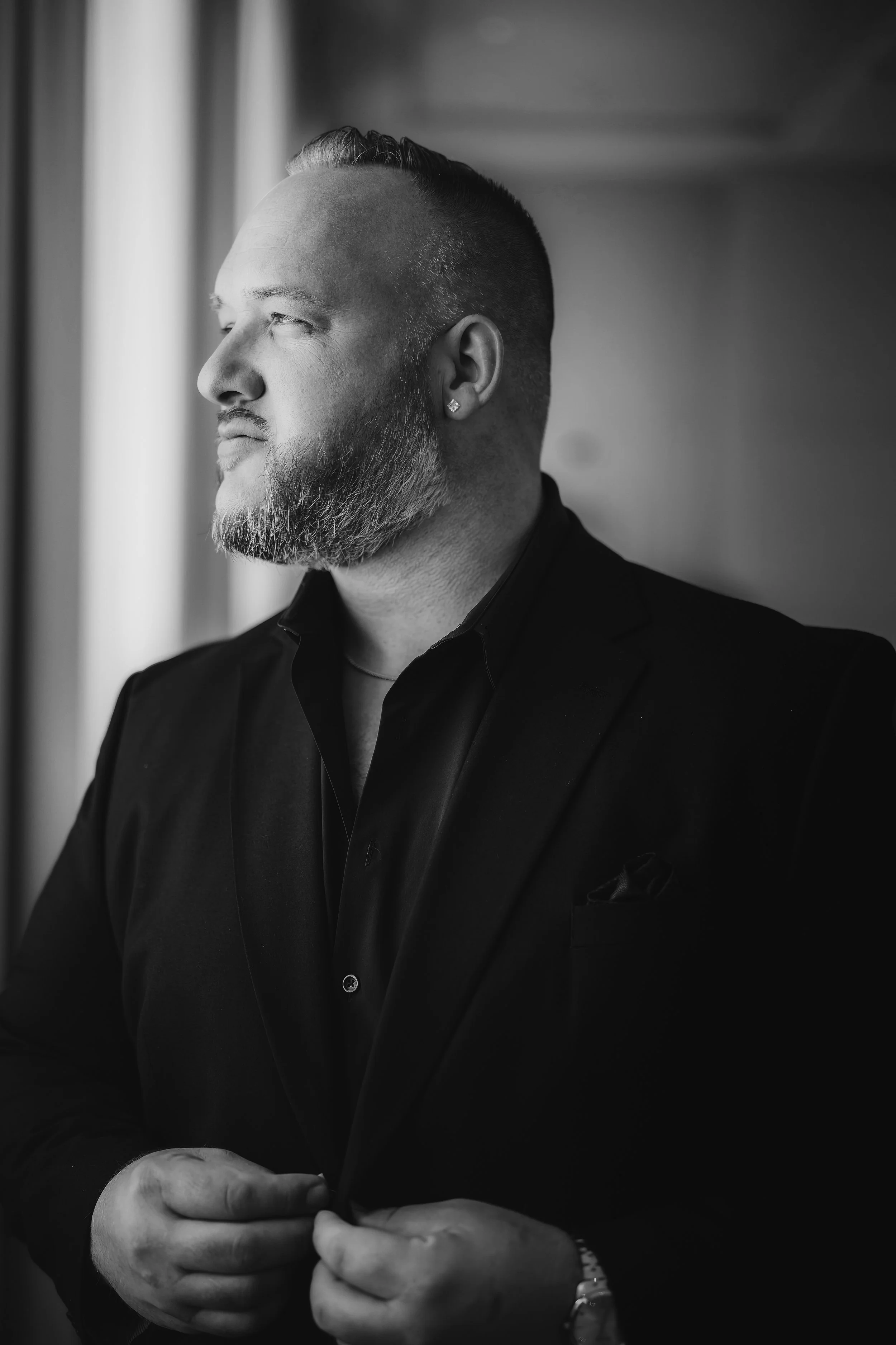 A black and white photo of a man with a beard, wearing a suit and wristwatch, adjusting his shirt cuff while looking out a window.