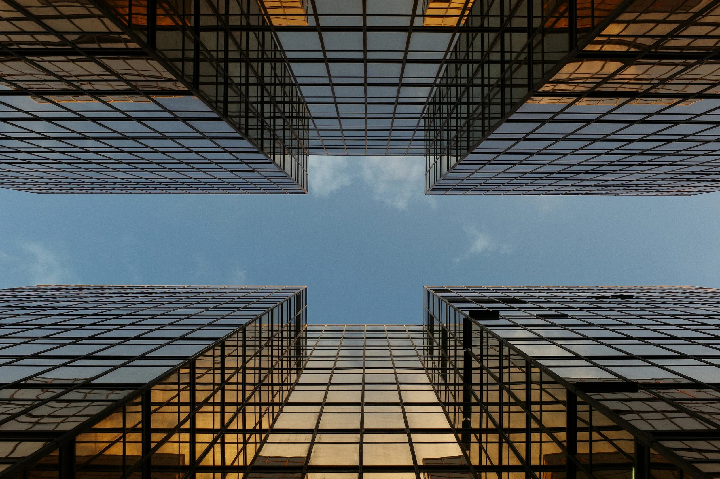 Looking up at the sky through the gaps between tall modern office buildings with glass facades.