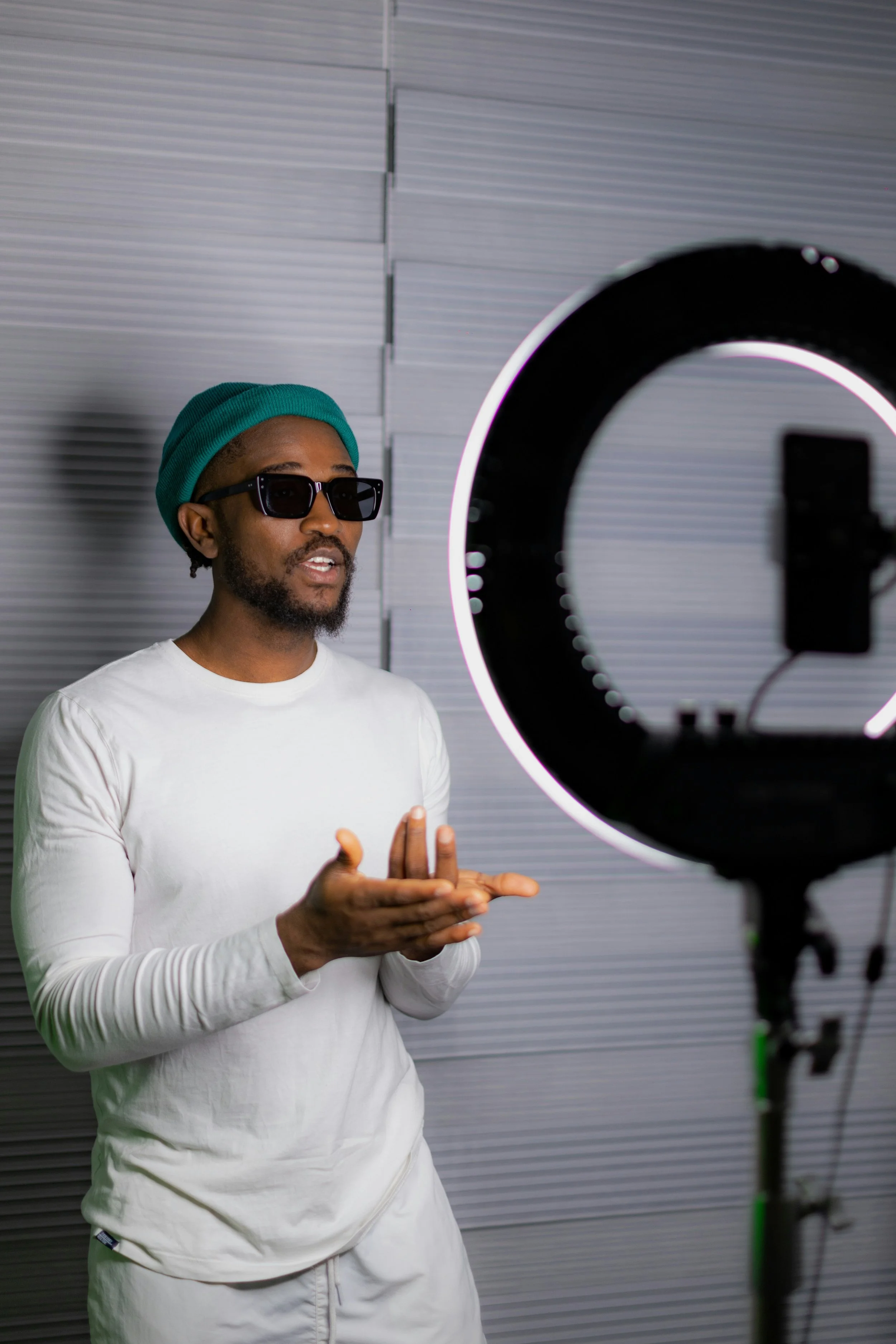 A man wearing sunglasses, a white long-sleeve shirt, white pants, and a teal beanie, facing a ring light and gesturing with his hands.
