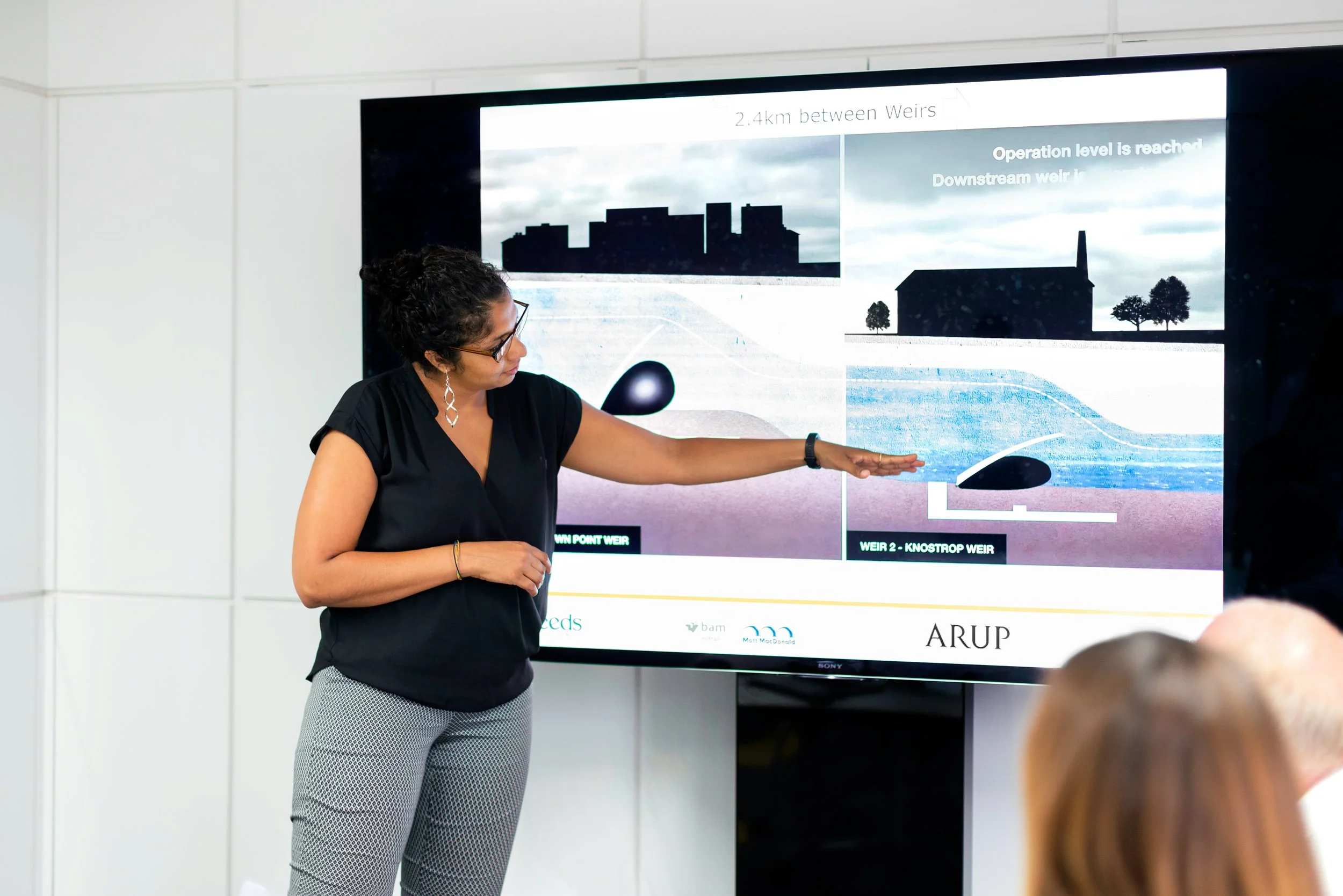 A woman in black and patterned pants, wearing glasses and earrings, points at a large screen displaying a presentation about underground water management with diagrams of weirs and underground structures.