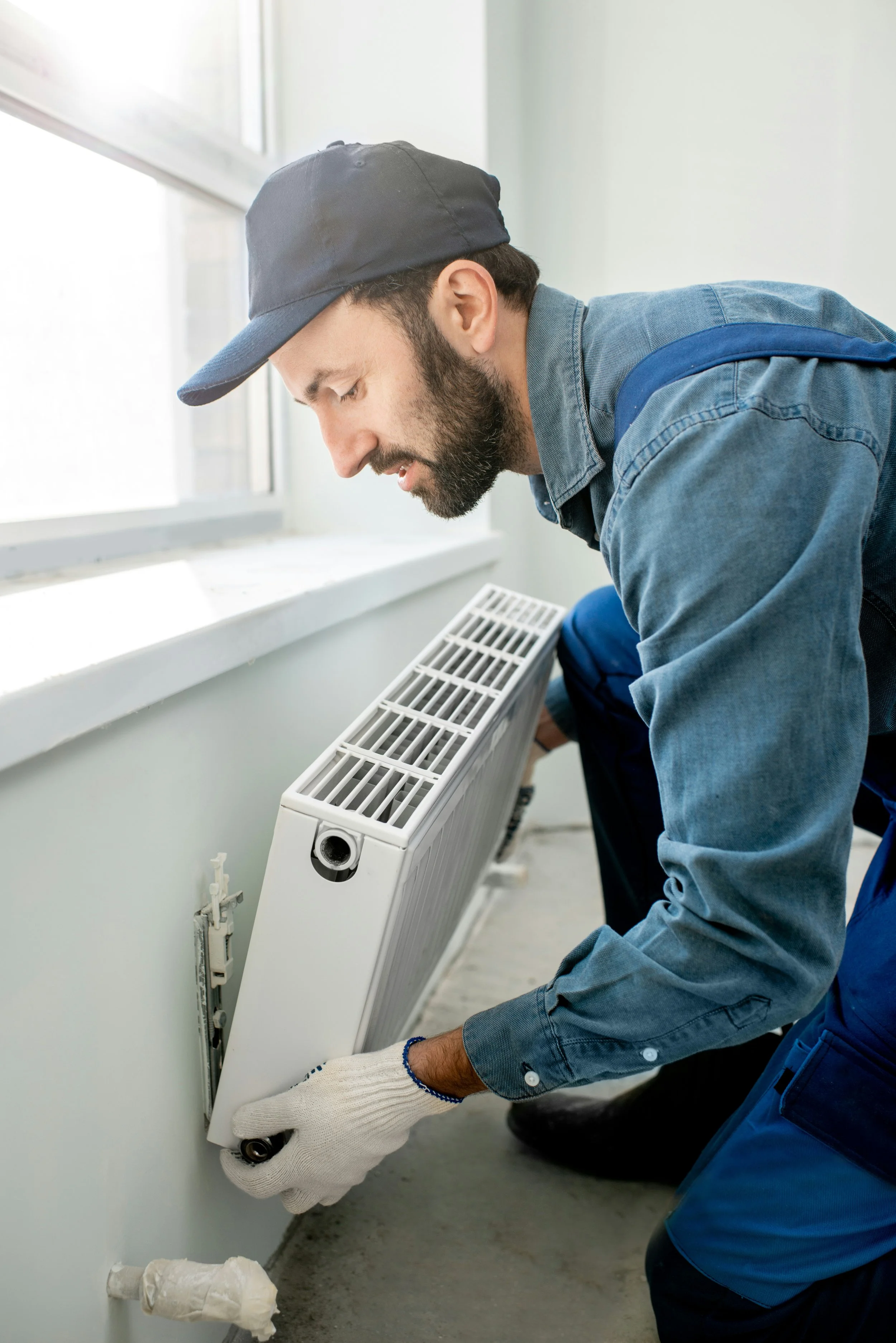 A man wearing a black cap, denim shirt, and gloves installing or repairing a white radiator near a window.