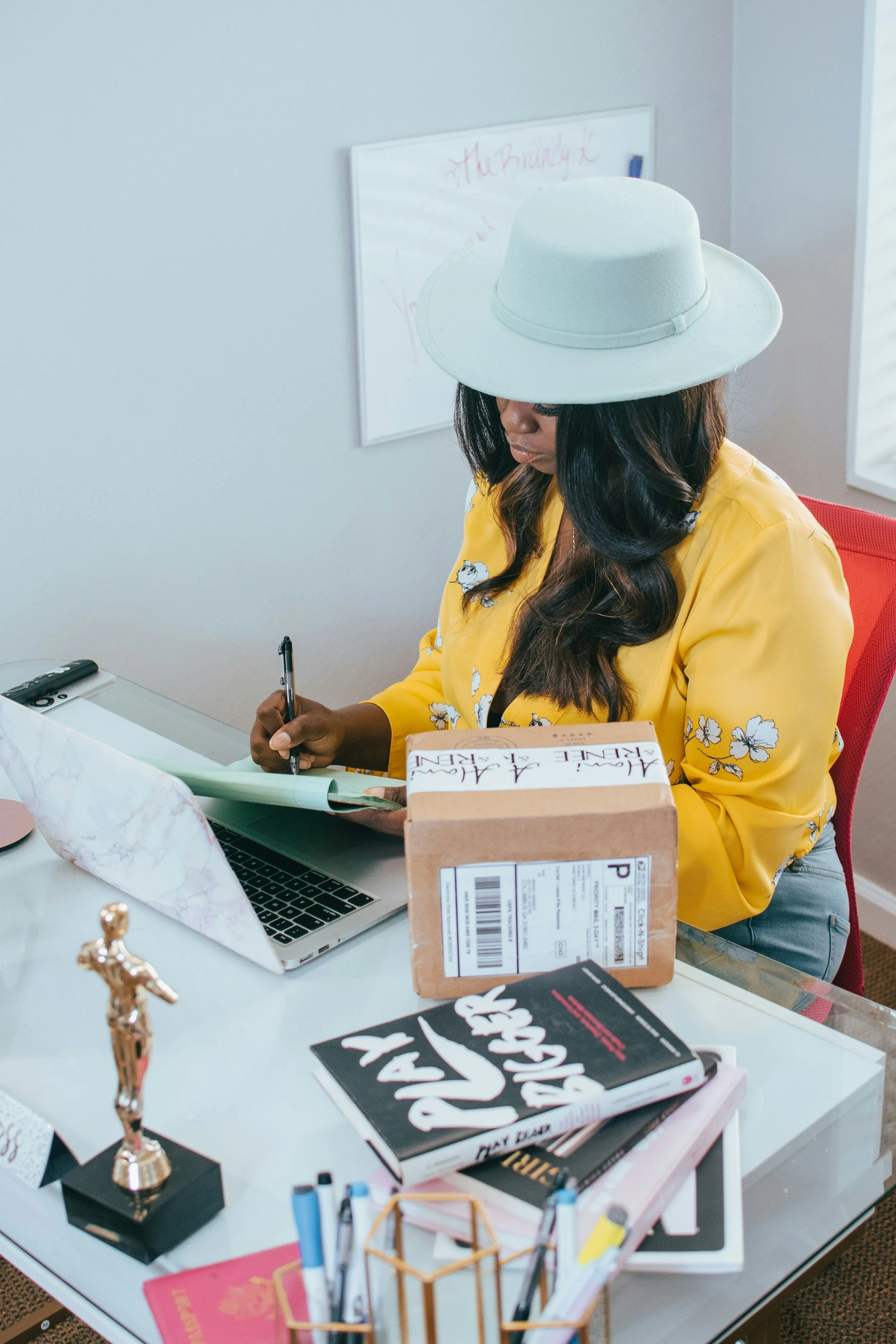 Woman with long black hair wearing a large white hat and yellow floral shirt sitting at a white desk, writing on a notepad, with a laptop, a medium-sized package, a black book, a pink notebook, and various stationery items in the foreground.