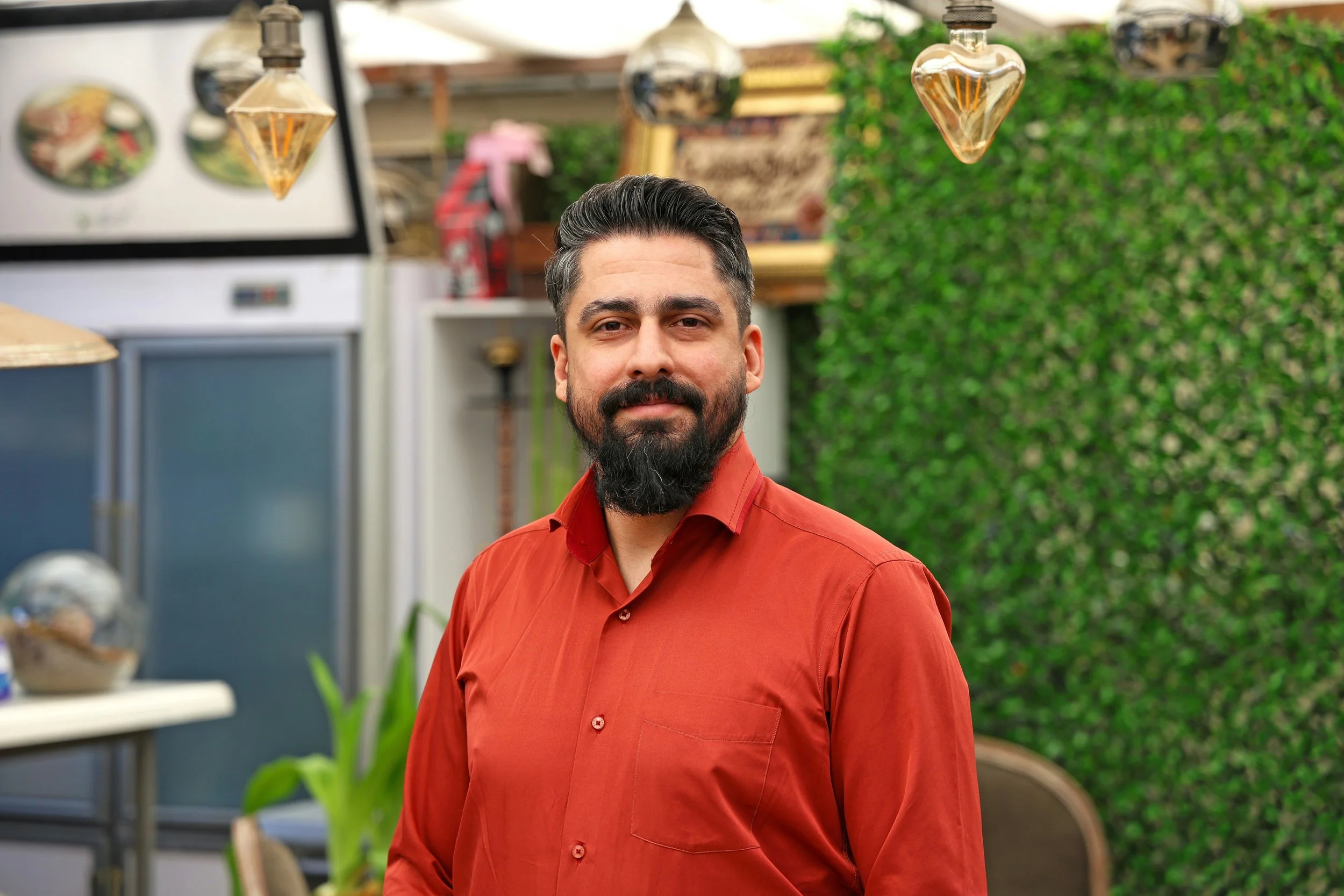 A man with dark hair, a well-groomed beard, wearing a red shirt, standing in a greenery-filled outdoor space with hanging glass lights and plants in the background.