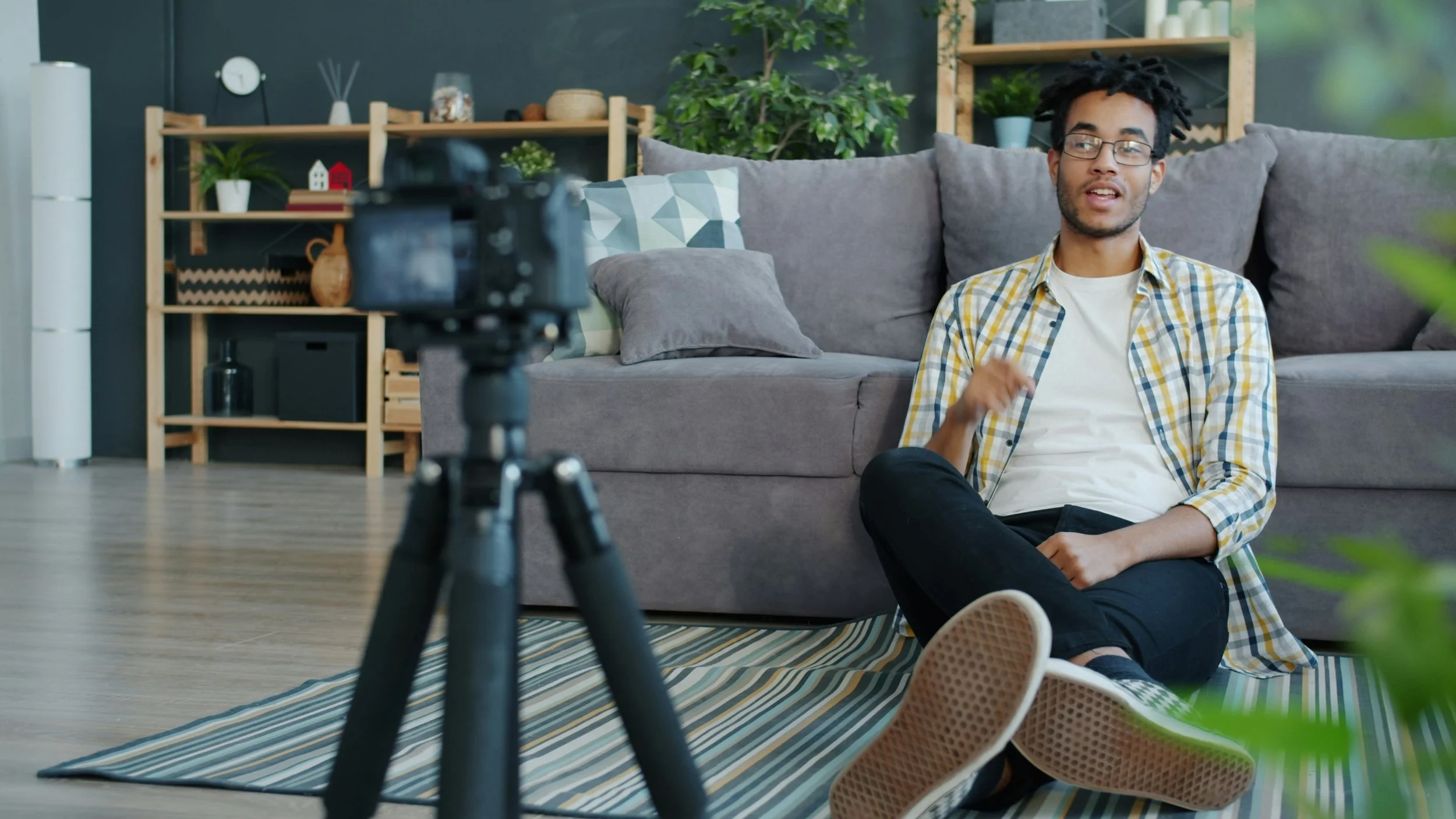 A young man with glasses and dreadlocks sitting on the floor in front of a camera, recording a video in a living room with a gray sofa and wooden shelves filled with decorative items and plants.