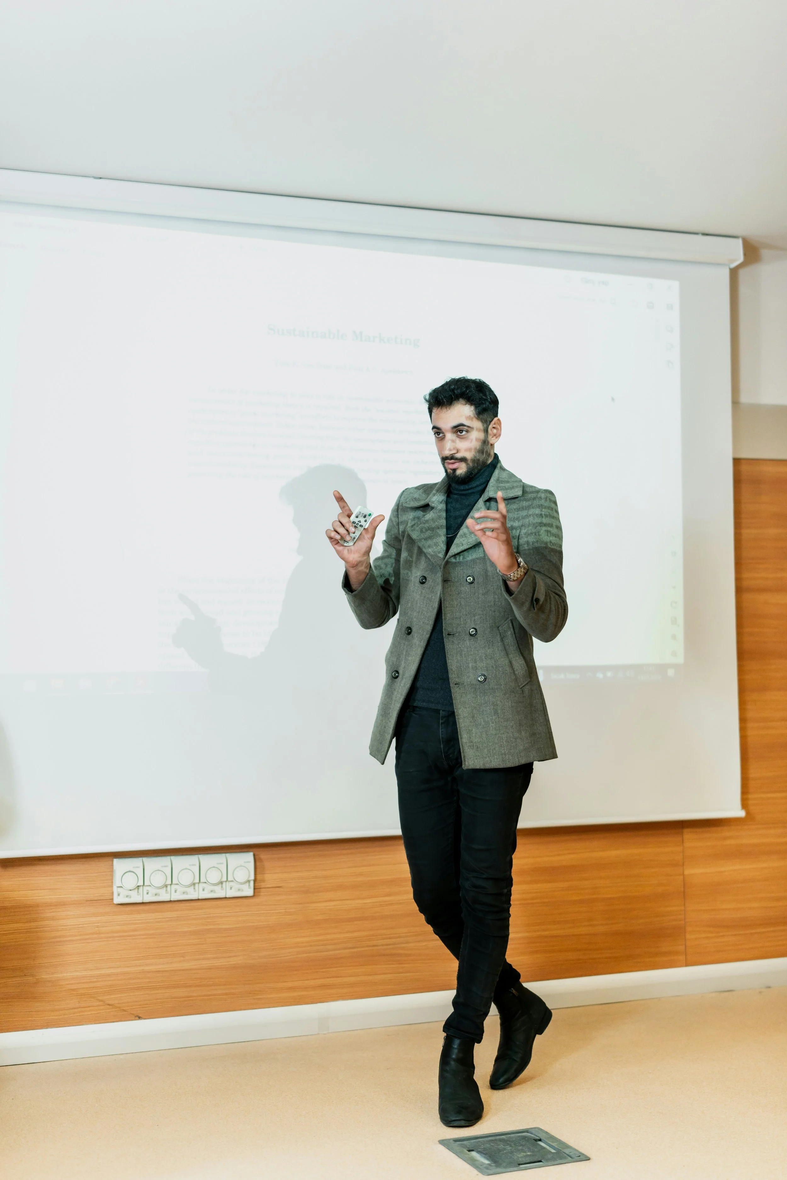 A man standing in front of a presentation screen, holding a remote control, dressed in a coat, black pants, and boots, with a presentation on sustainable marketing projected on the screen behind him.