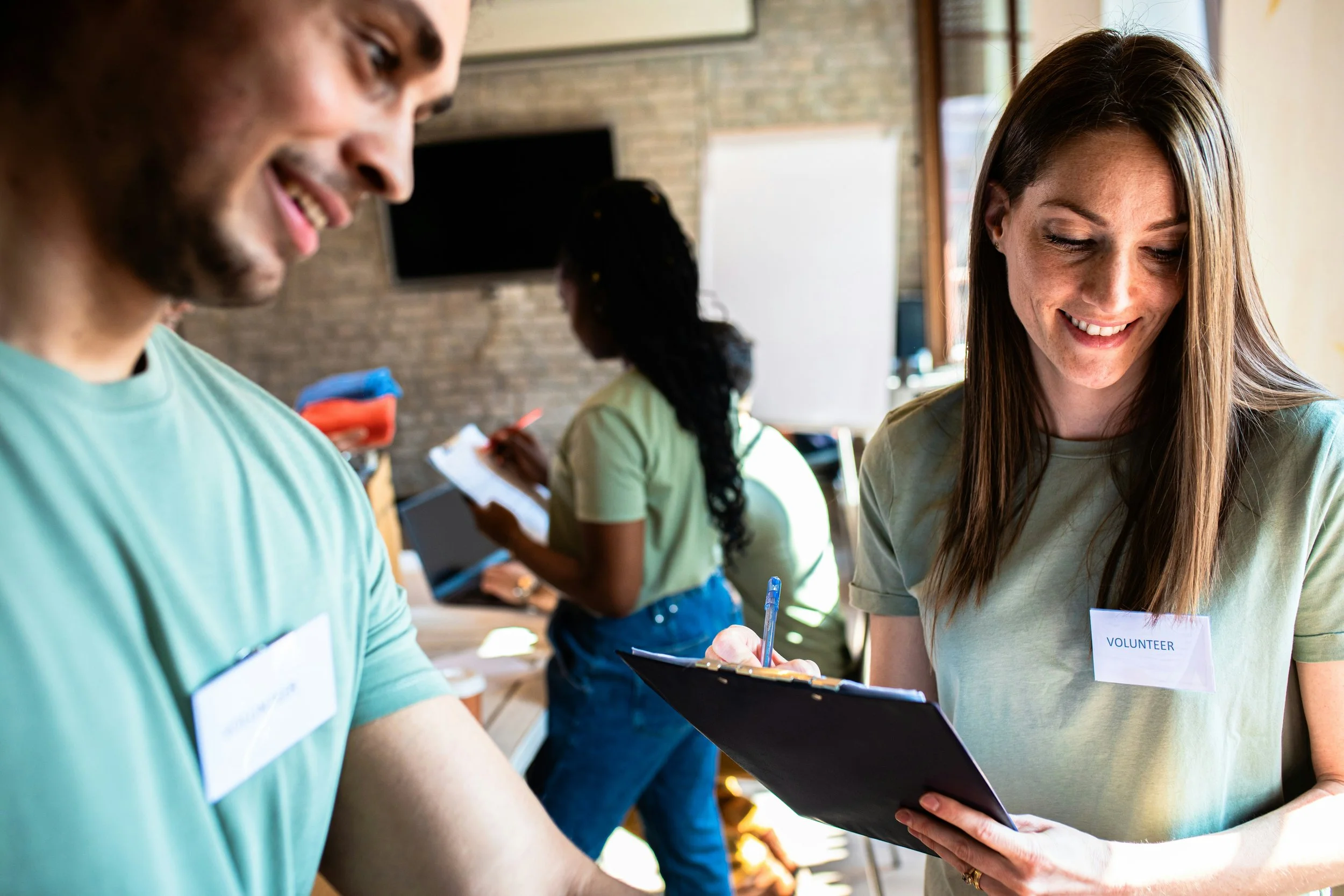 Smile of a young woman wearing a volunteer badge signing a form while standing next to a man in a green shirt with a volunteer badge, in a room with brick walls and large windows.