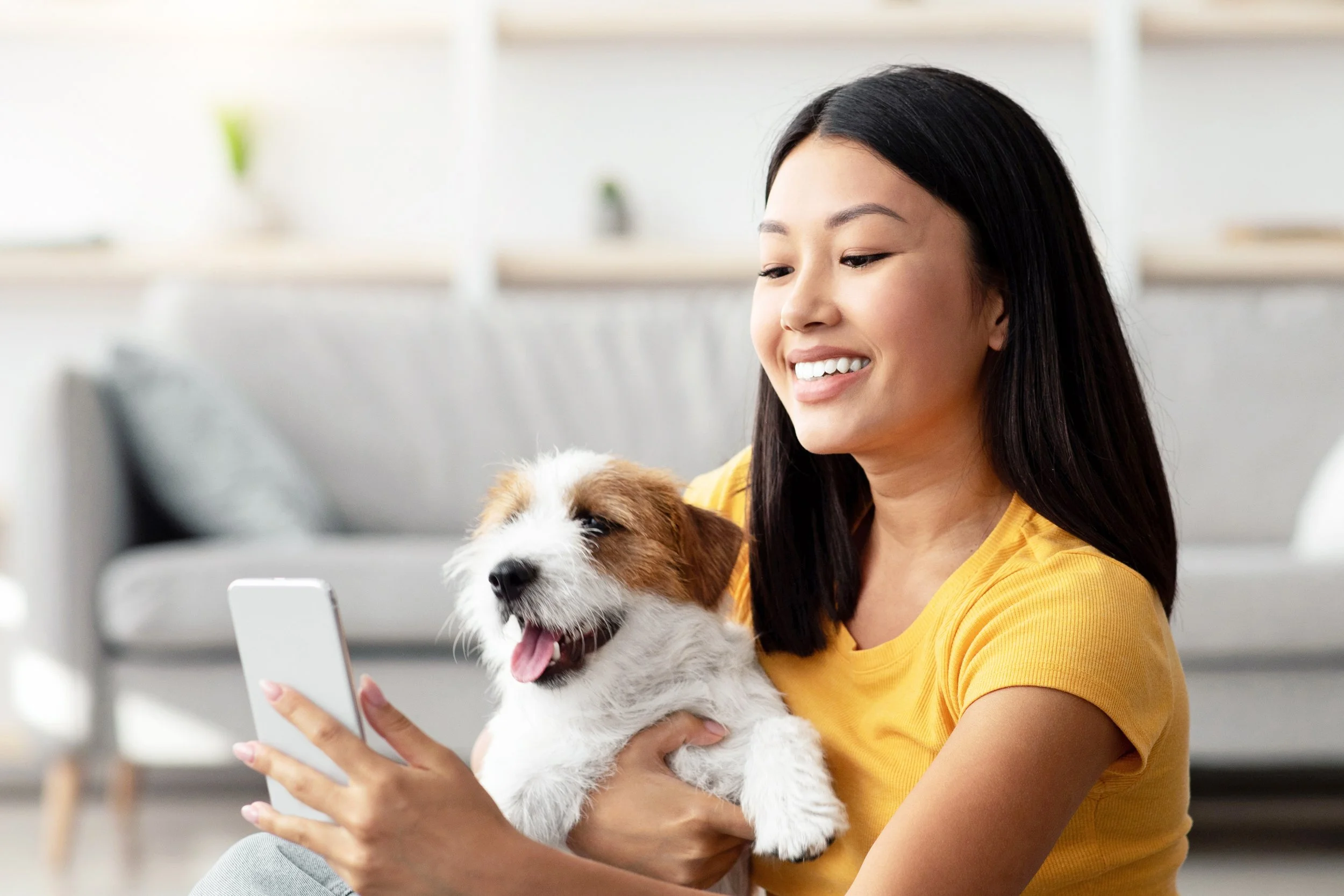 Woman in yellow shirt holding her dog is looking into her phone and smiling.
