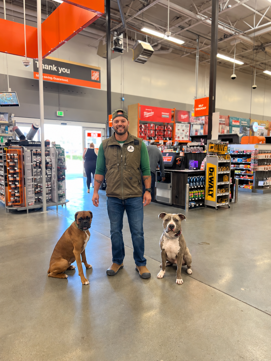 A man with two dogs inside a hardware store near checkout counters. The man is smiling, wearing a green shirt, a vest, jeans, and a cap backward. The dogs are sitting on the floor, one is brown with a black face and the other is gray and white, both looks alert.