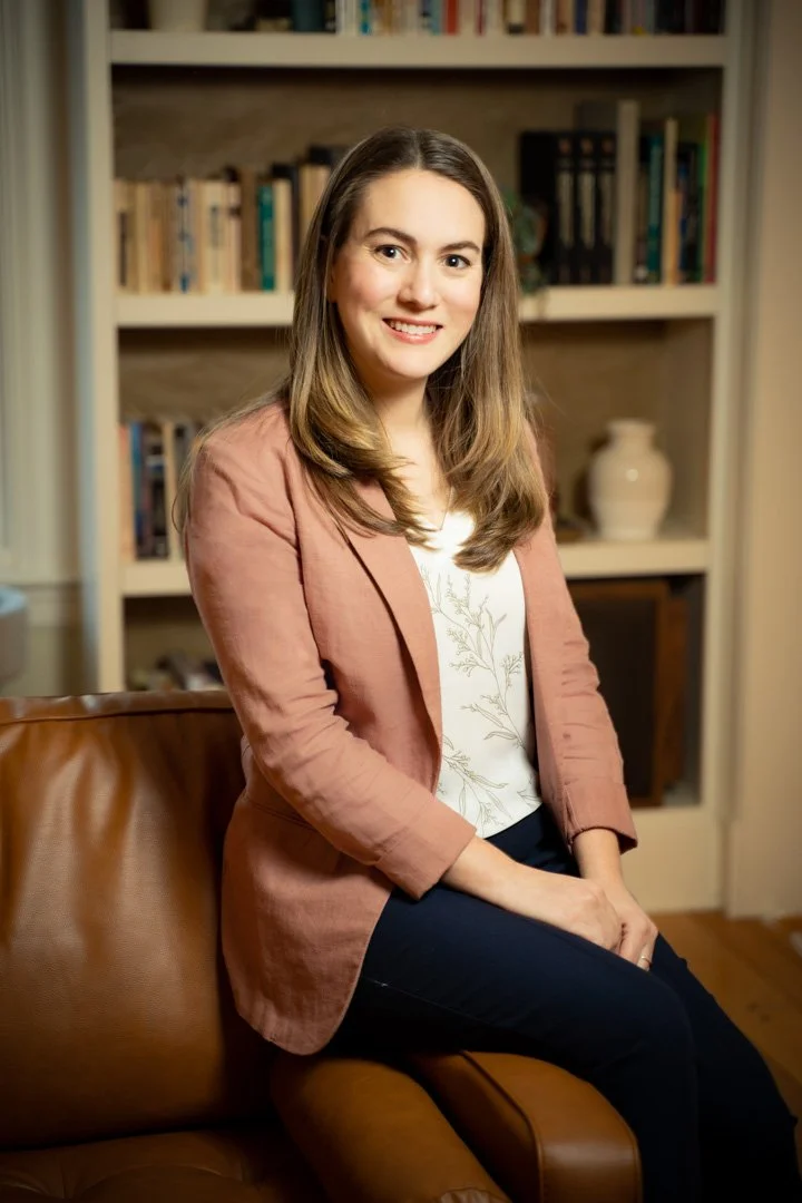 A woman with long brown hair, wearing a pink blazer and dark pants, sitting on a brown leather couch in a room with a bookshelf filled with books and decorative items in the background.