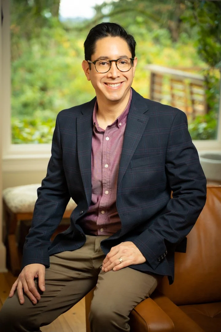 A man with short black hair and glasses smiling while sitting indoors near a window with greenery outside. He is wearing a dark blazer, a maroon button-up shirt, and khaki pants.