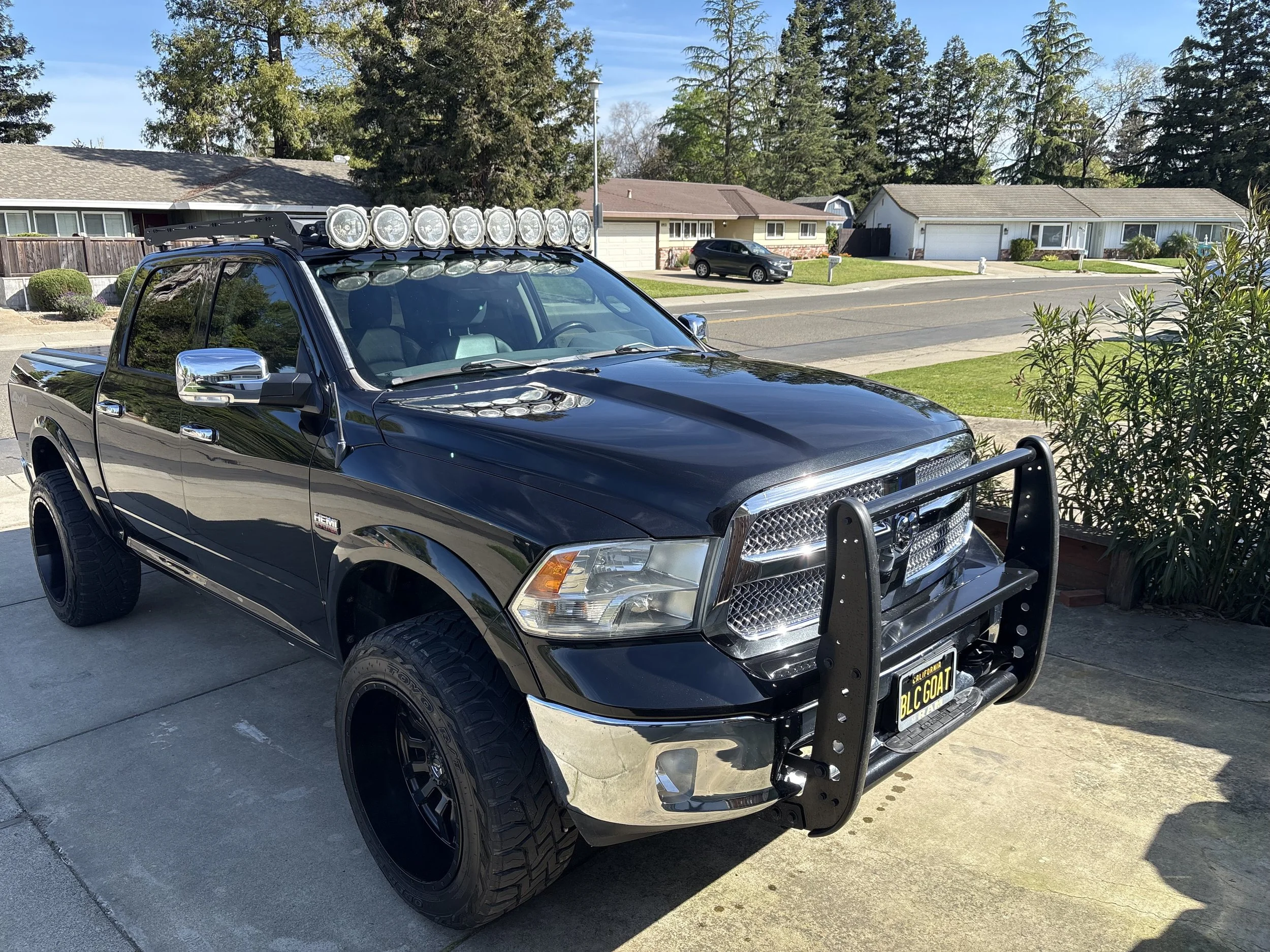 A black pickup truck with off-road tires and a front bumper guard parked in a residential driveway. It has a large roof rack with multiple bright headlights mounted on top and a California license plate reading 'BLC GOAT.' In the background, there are single-story houses with well-maintained lawns and a street lined with trees under a clear blue sky.