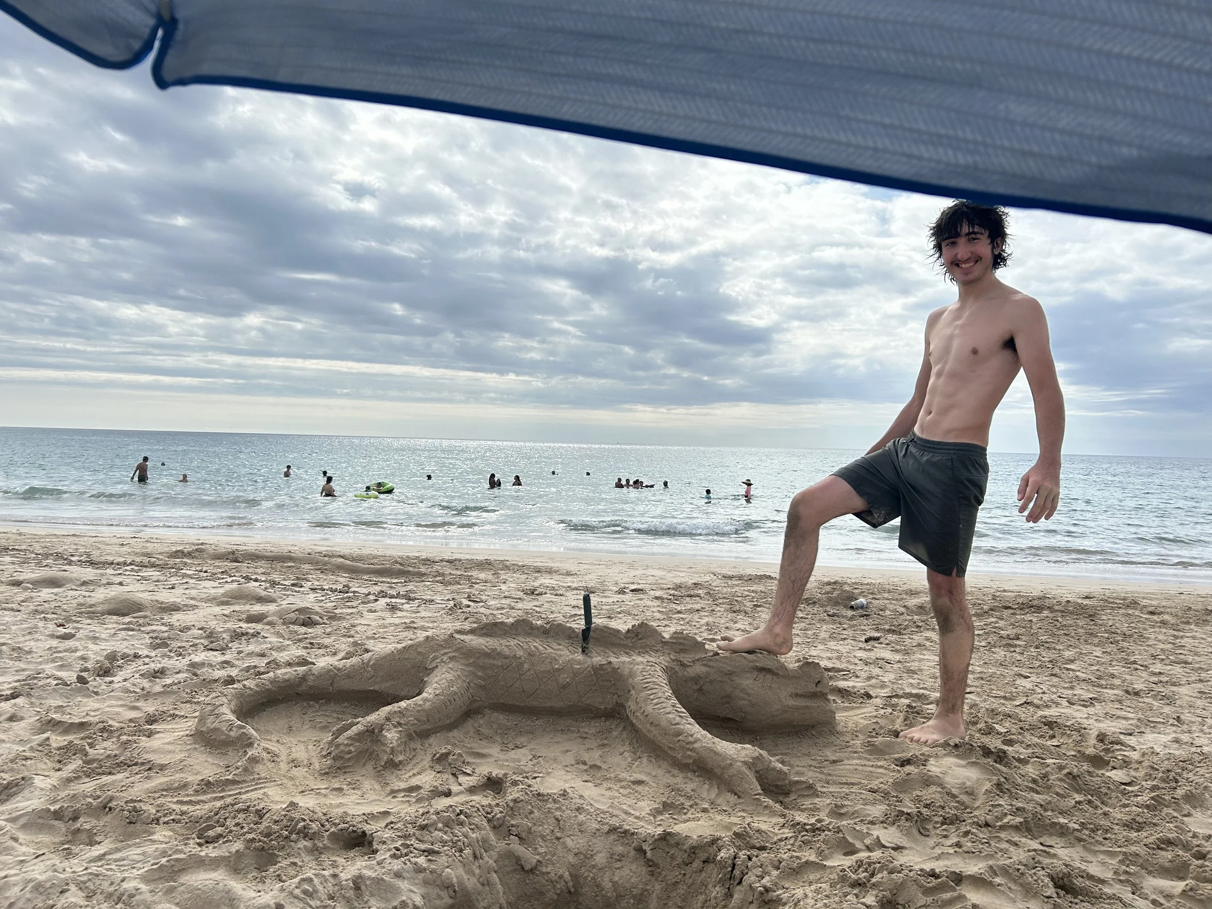 A young man with dark, curly hair and a mustache is standing on a sandy beach, smiling at the camera. He is shirtless and wearing dark swim trunks, with one foot slightly raised. In the background, there are several people in the water, some with floatation devices, and the sky is cloudy over the ocean.
