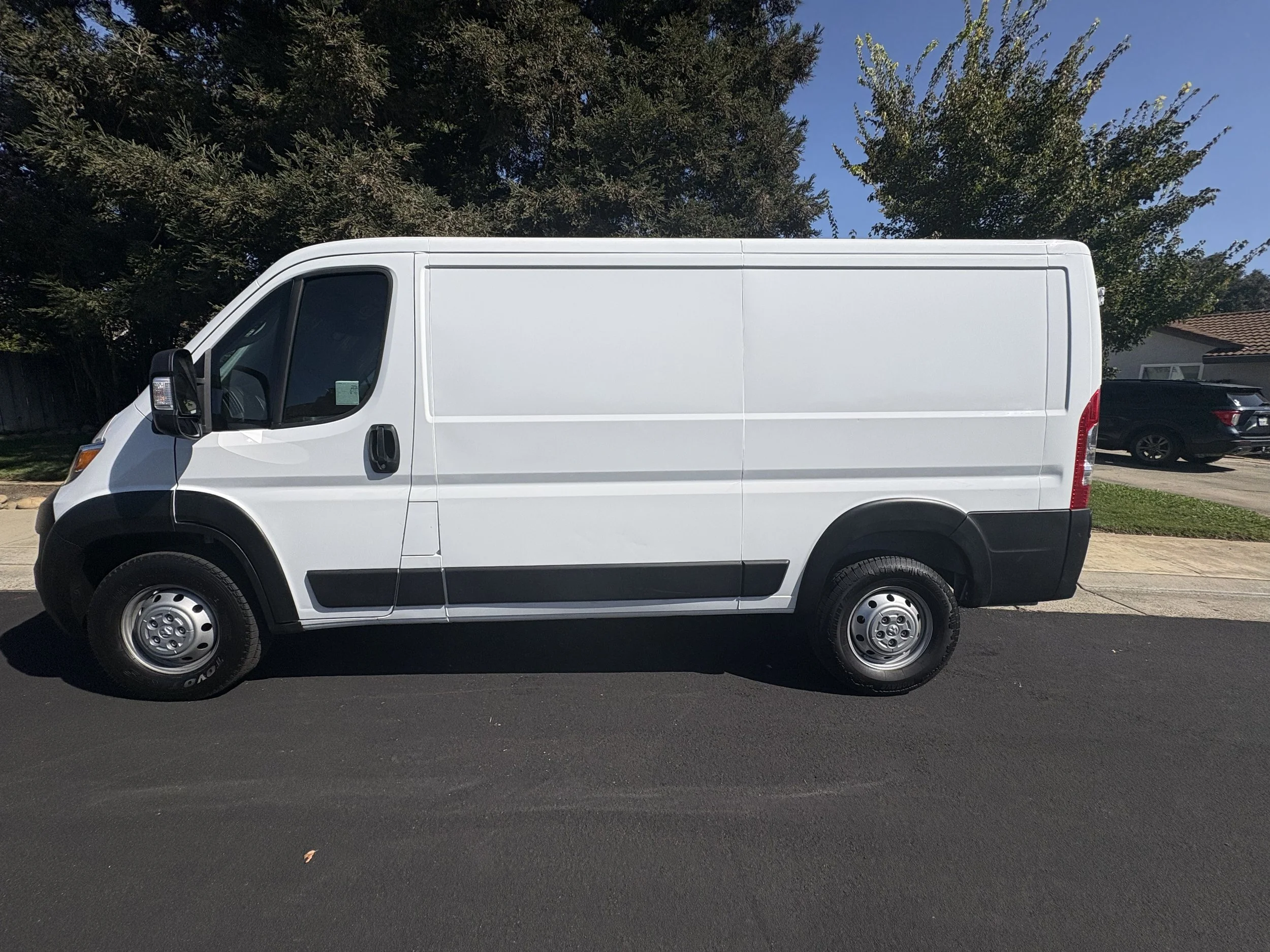 White cargo van parked on a residential street with a house and large tree in the background.